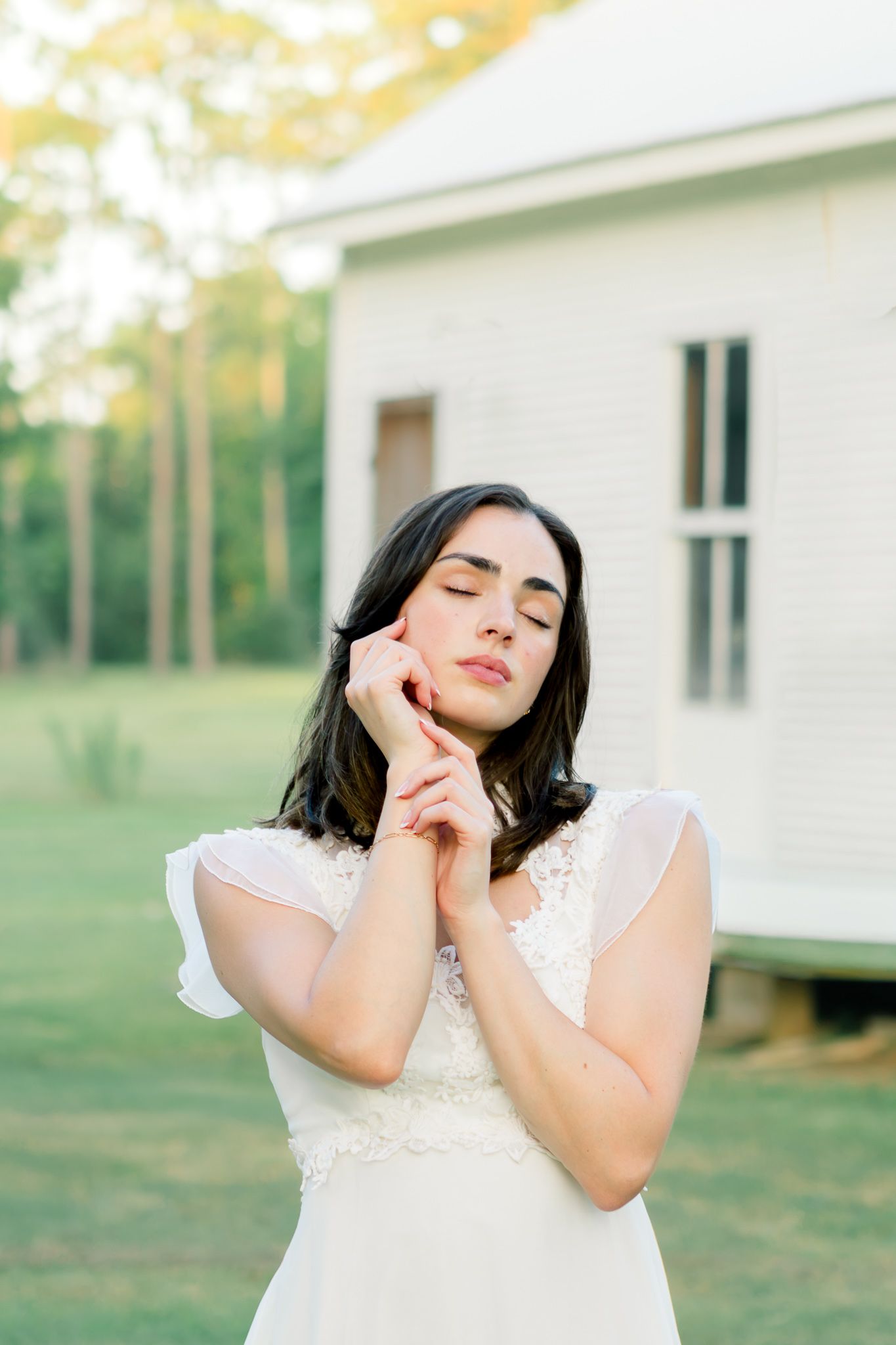 A bride in a white lace dress poses with closed eyes near a white chapel surrounded by greenery.