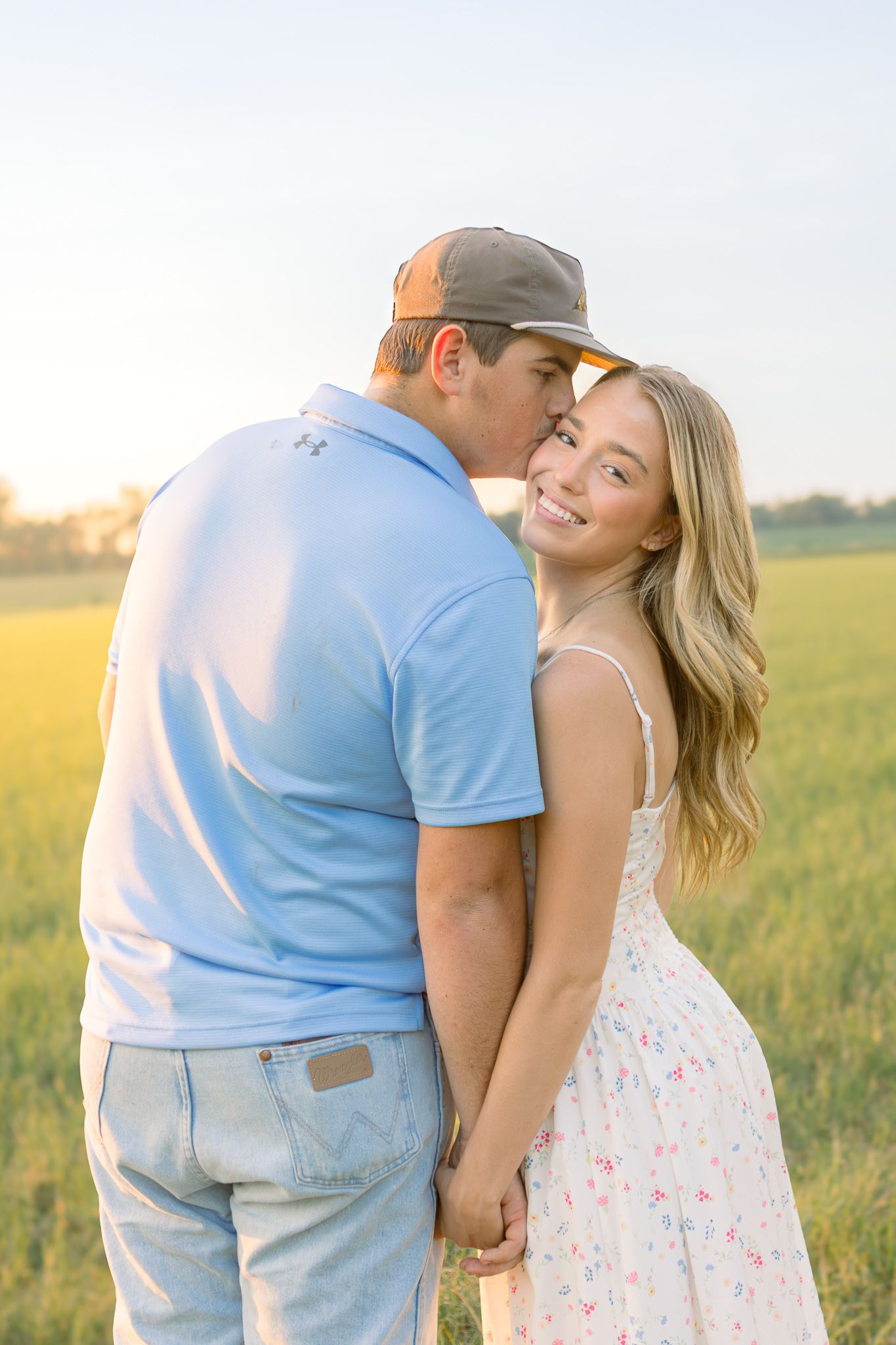 A couple holds hands in a golden field at sunset, the man kissing the smiling blonde woman on the cheek.
