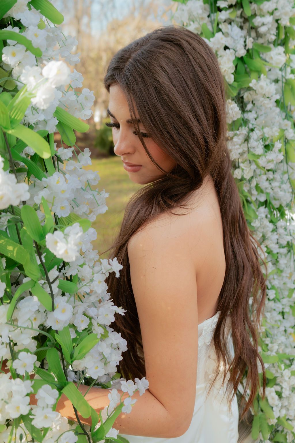 Brunette bride in a white strapless dress surrounded by white blossoming flowers in an outdoor setting.