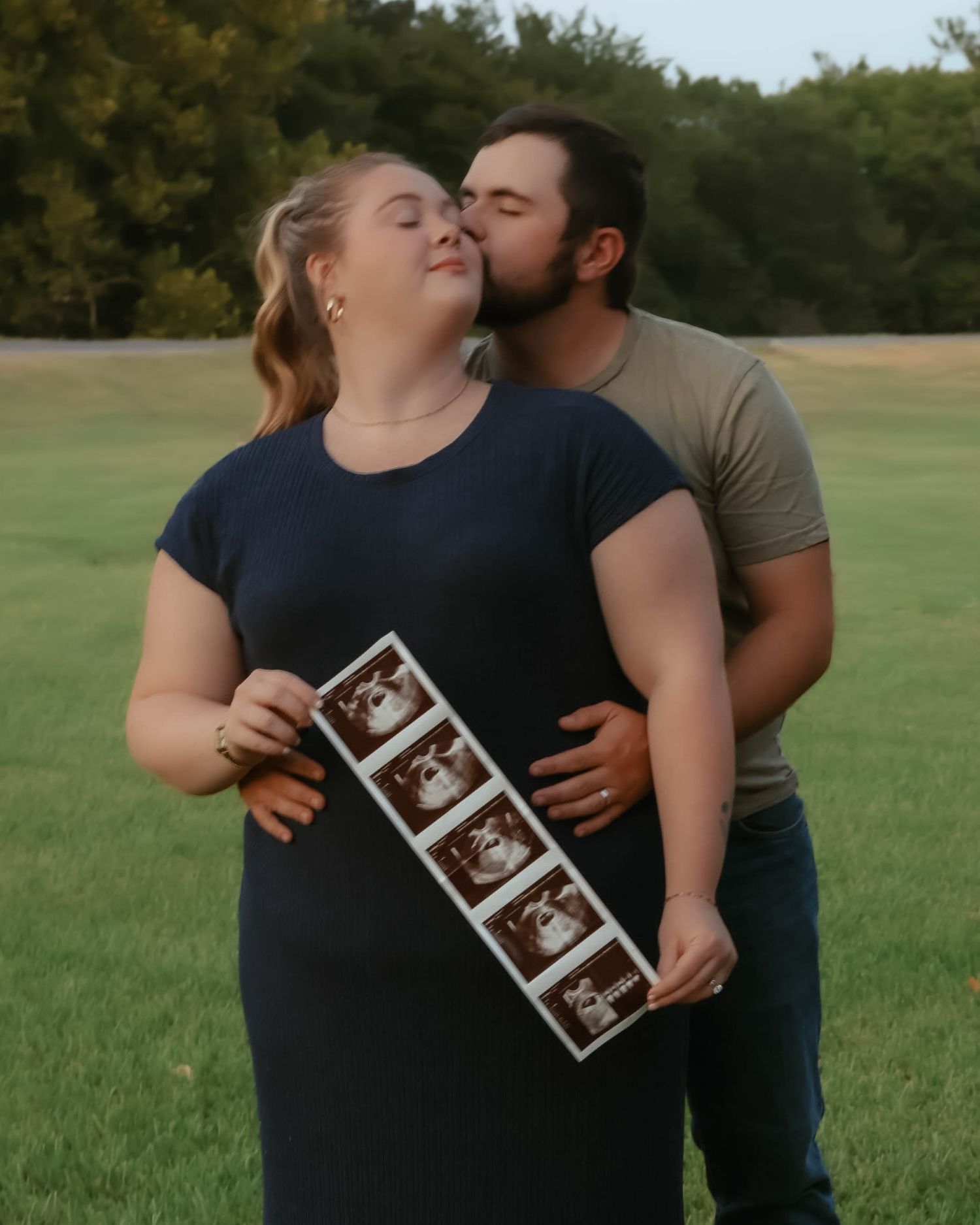 Couple sharing an intimate moment in a grassy field while holding ultrasound photos.