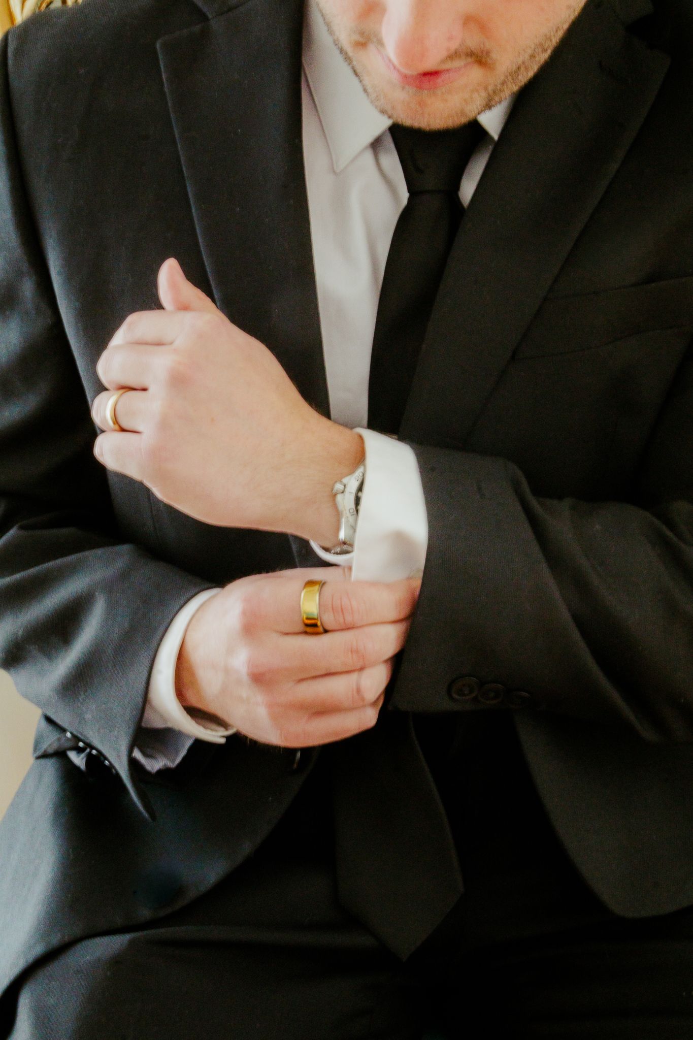 Groom in black suit adjusting cufflinks with gold wedding band and ring visible on fingers.