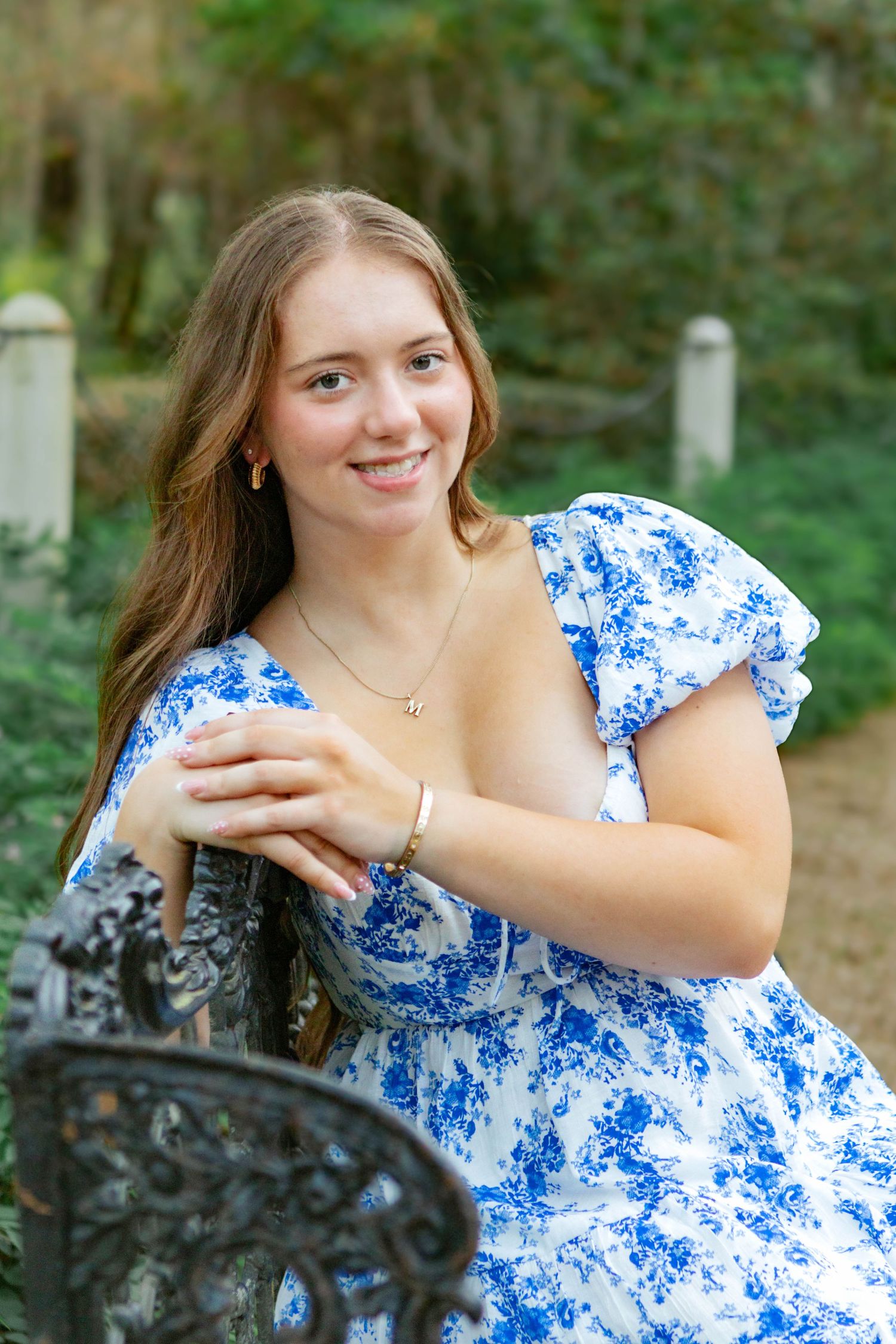 Young woman in blue floral dress leaning on ornate iron chair smiling outdoors in garden setting.