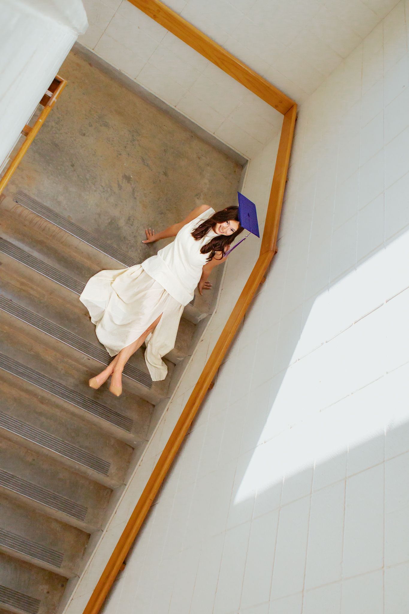 Overhead view of a young woman in white dress lying on wooden stairs holding a purple graduation cap.