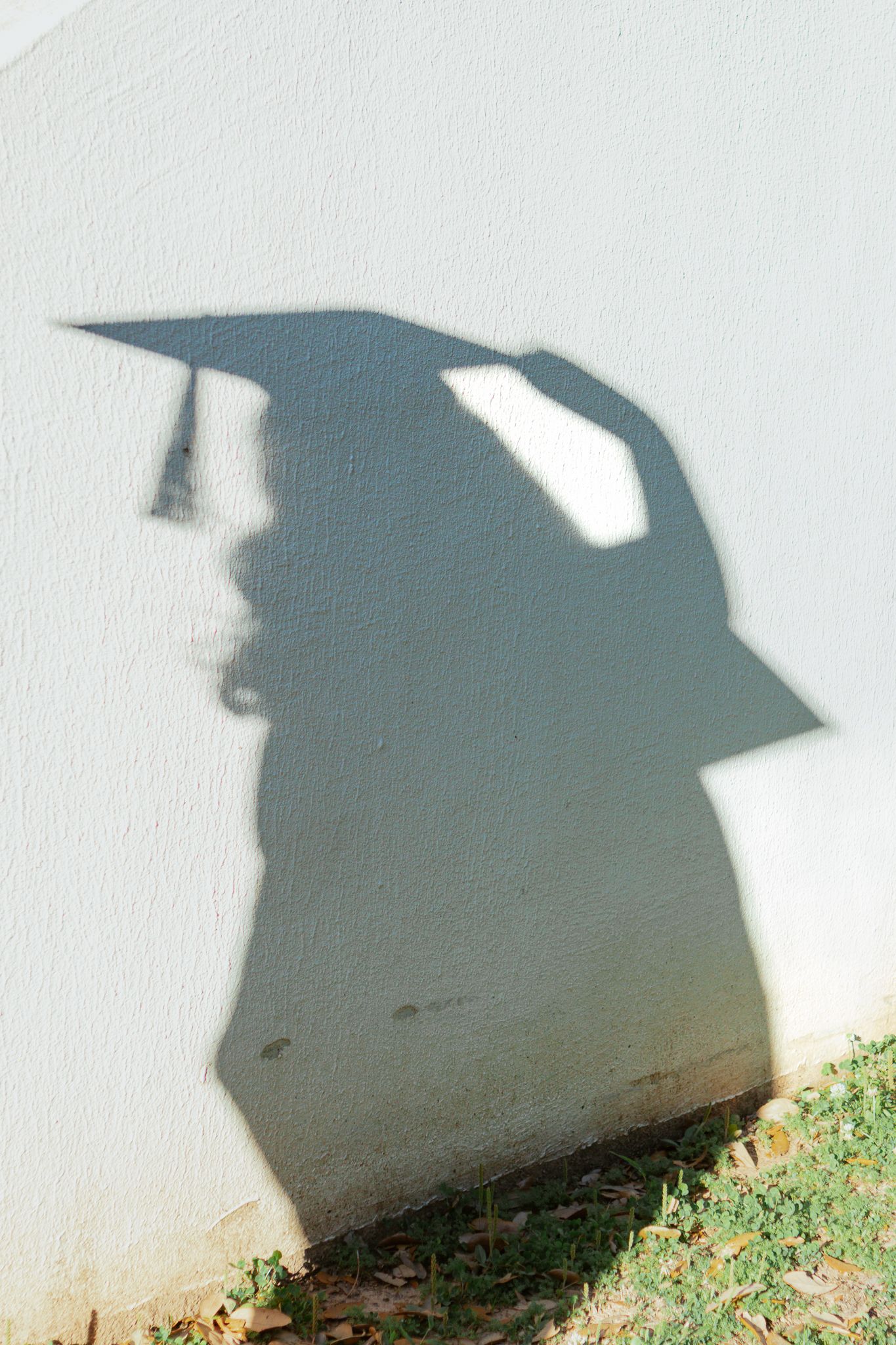 Shadow of a graduation cap and gown cast on a white textured wall with grass below.