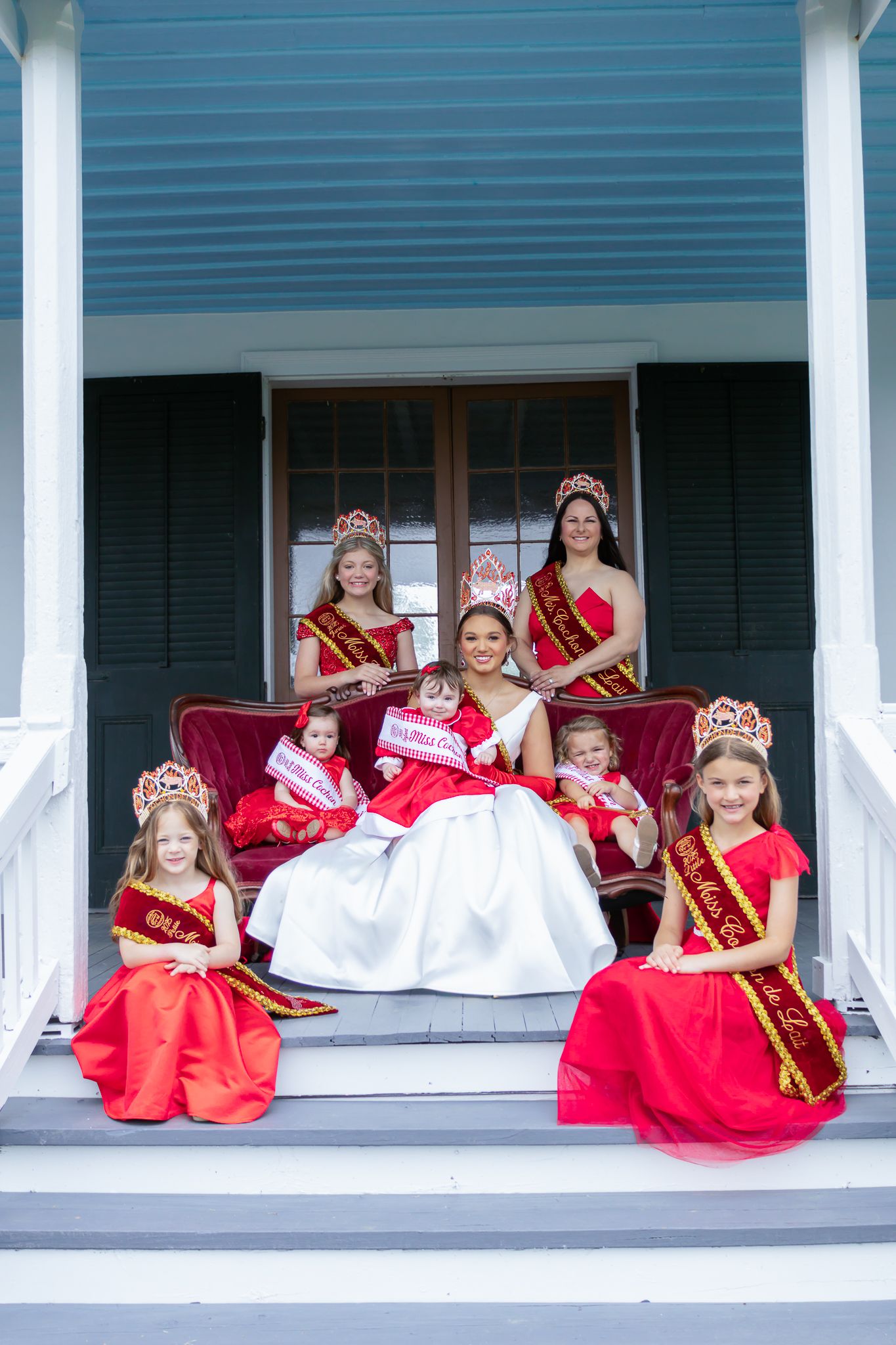 Beauty queen in white gown with court of red-dressed girls and crowns seated on porch steps.