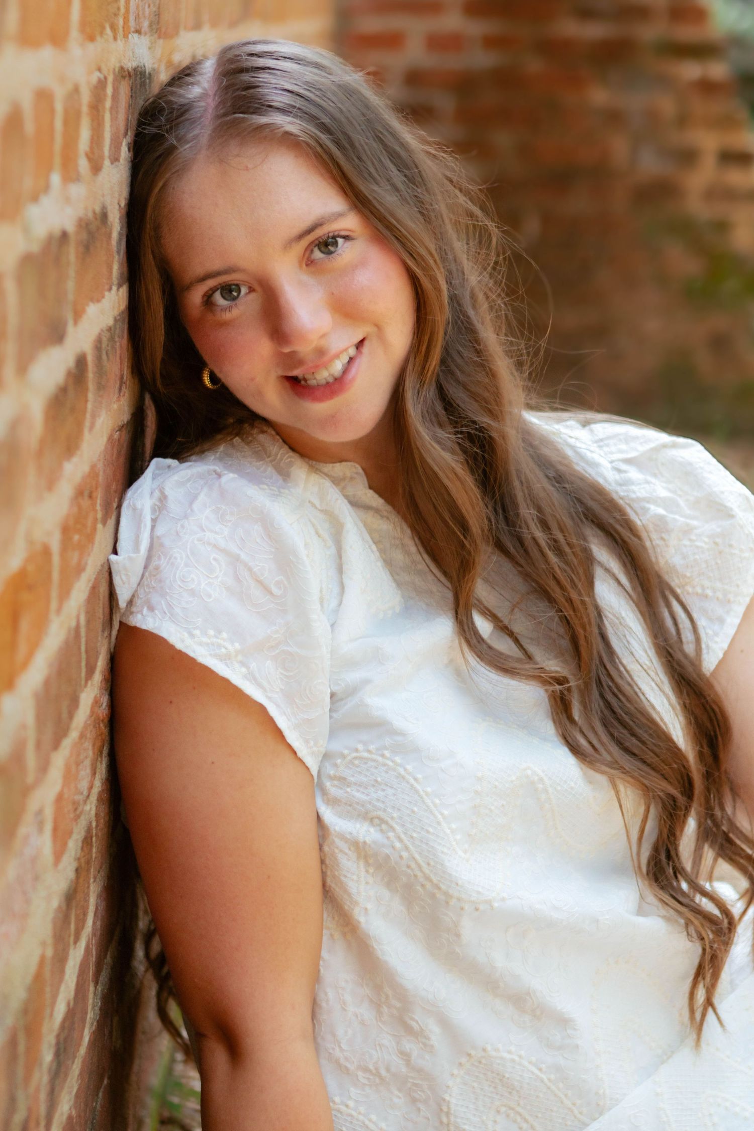 Smiling teenage girl with long brown hair in white lace top leaning against brick wall outdoors.