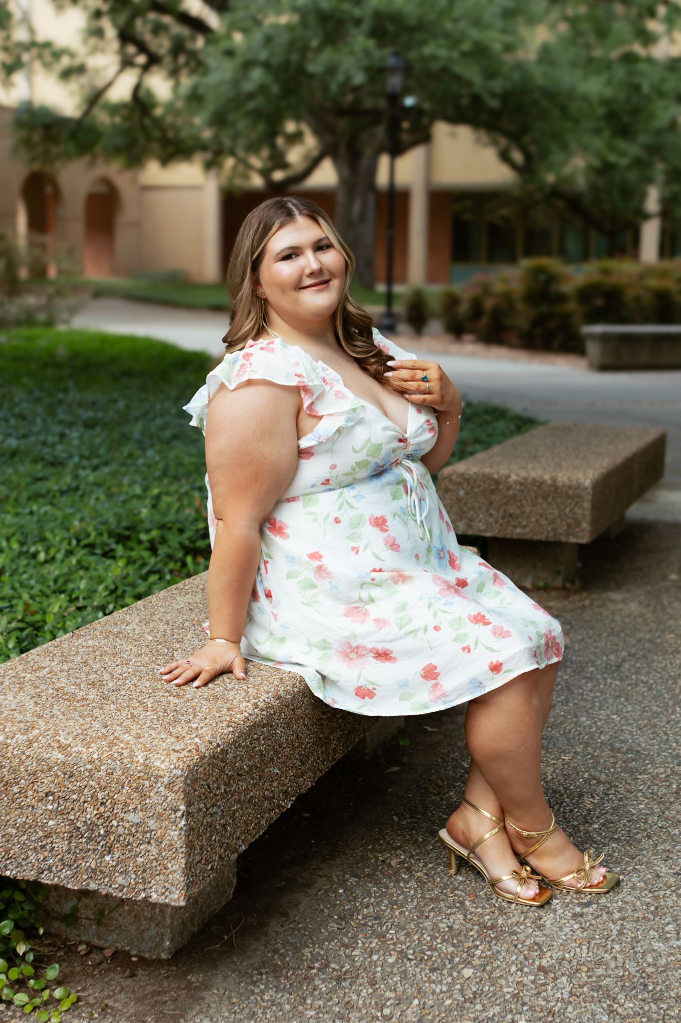 Young woman in floral sundress with gold heels sitting on a concrete bench in a landscaped courtyard.