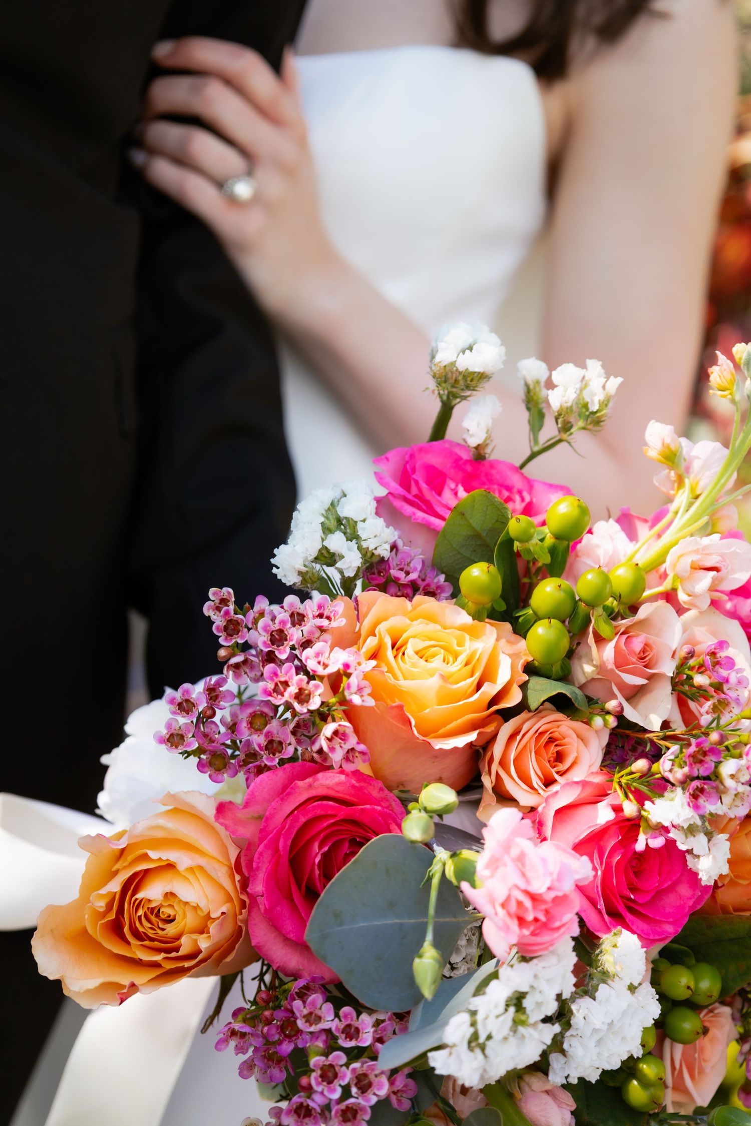 Bride and groom embracing behind a vibrant wedding bouquet of orange and pink roses with greenery.