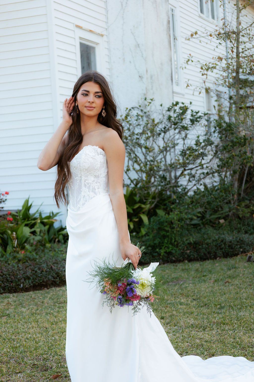 Bride in a strapless lace and satin gown holding a colorful wildflower bouquet outside a white historic building.