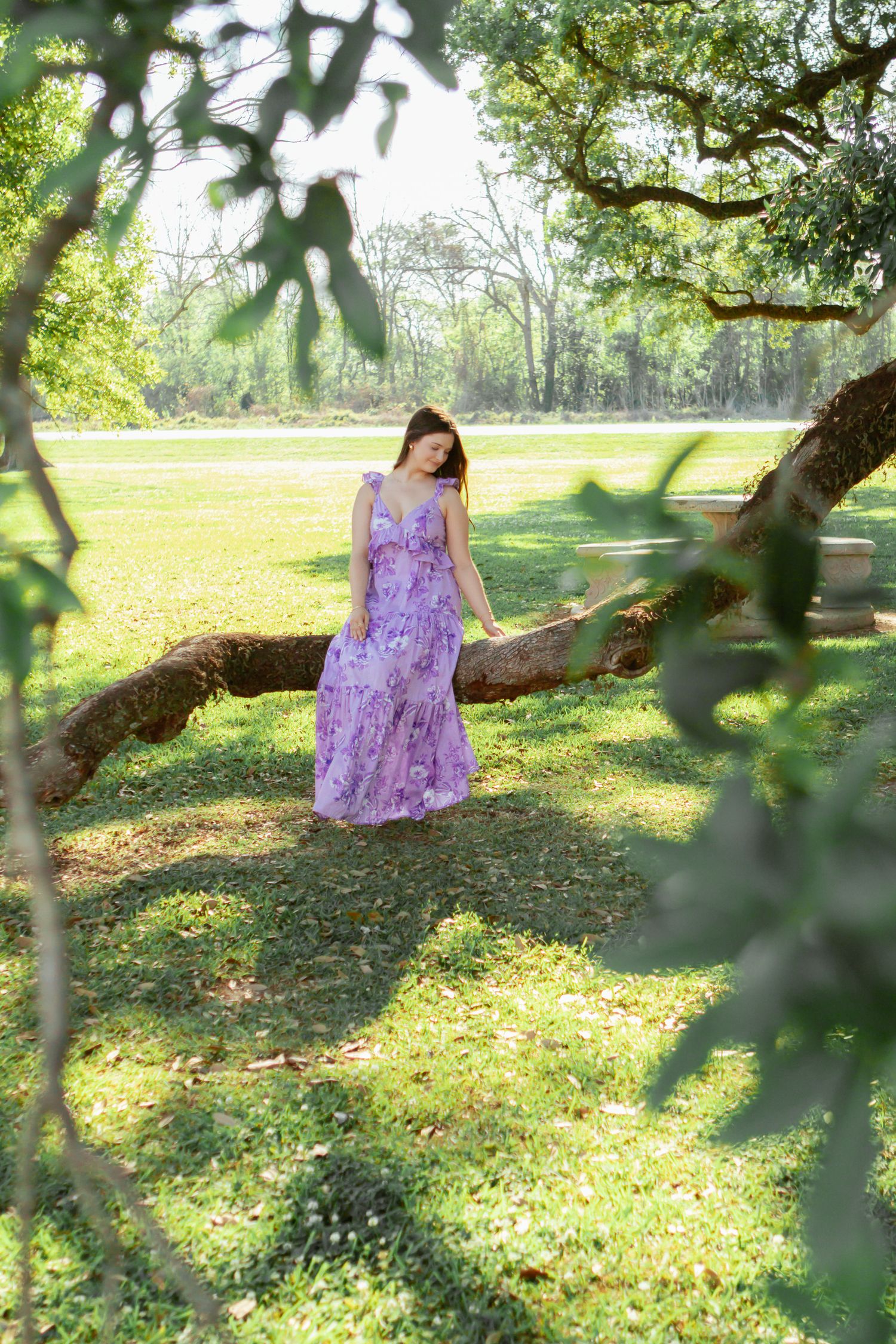 Young woman in purple floral maxi dress standing under large oak tree in lush green park.