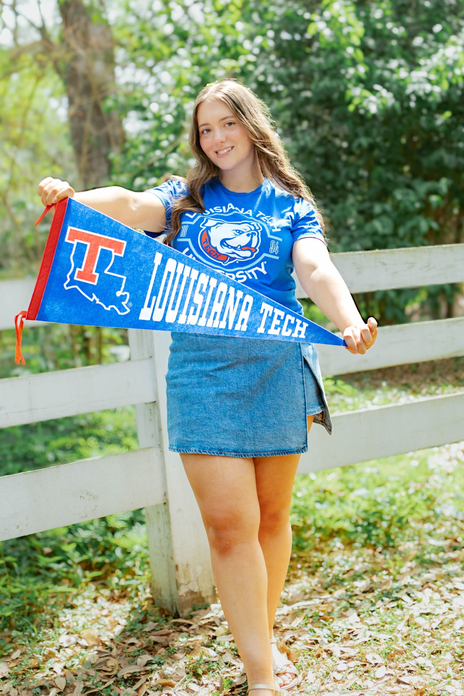 Young woman holding Louisiana Tech pennant wearing blue tie-dye shirt and denim skirt outdoors near white fence.