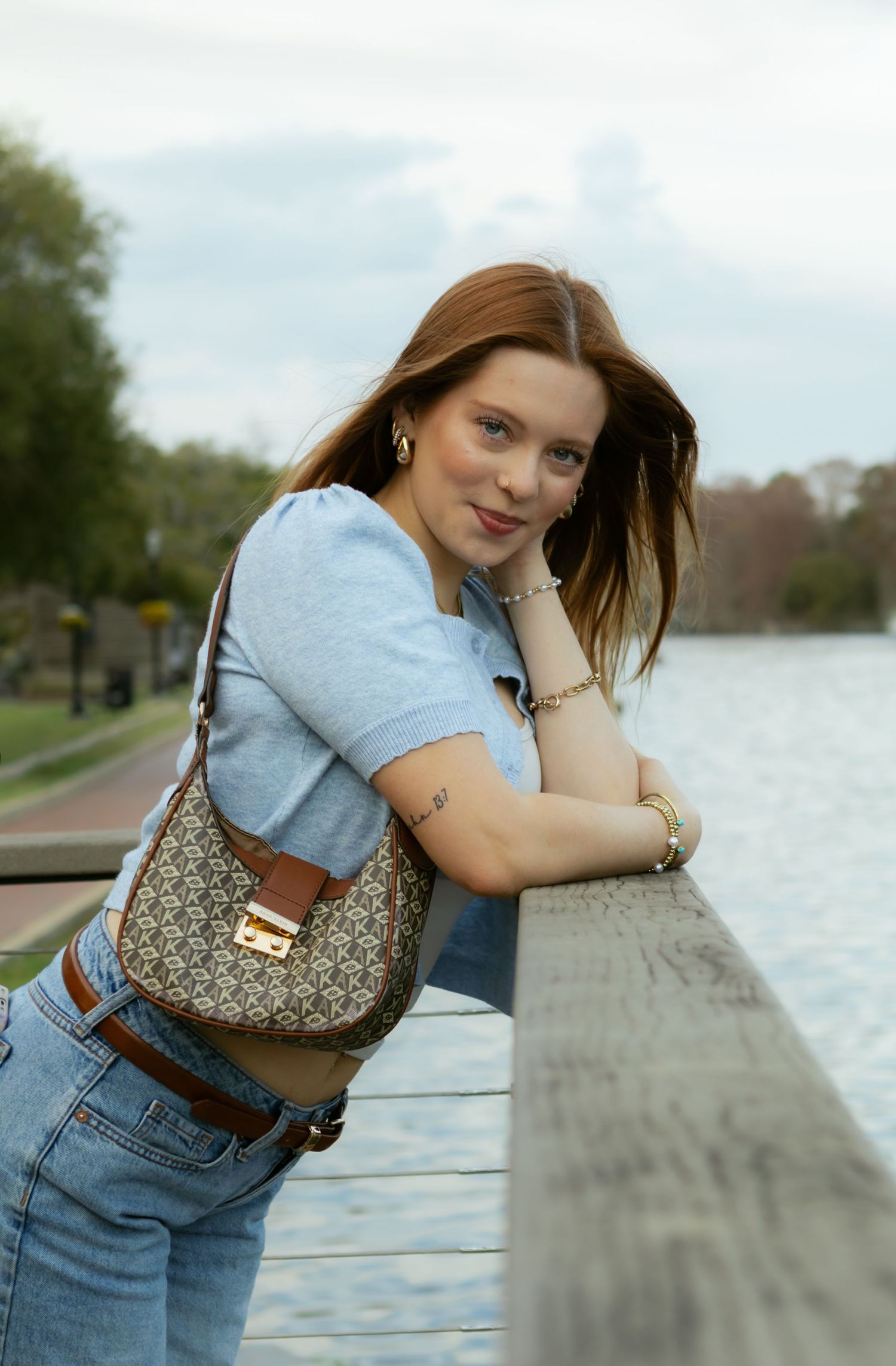 Young redhead woman leaning on a railing by a lake with a designer shoulder bag.