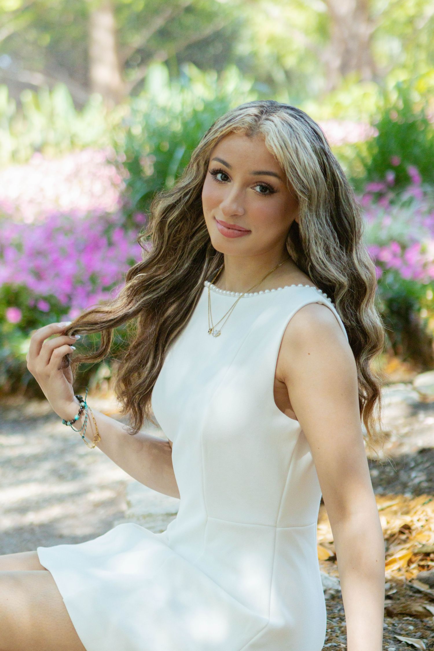 Young woman in white sleeveless dress sitting outdoors with pink flowers in background.