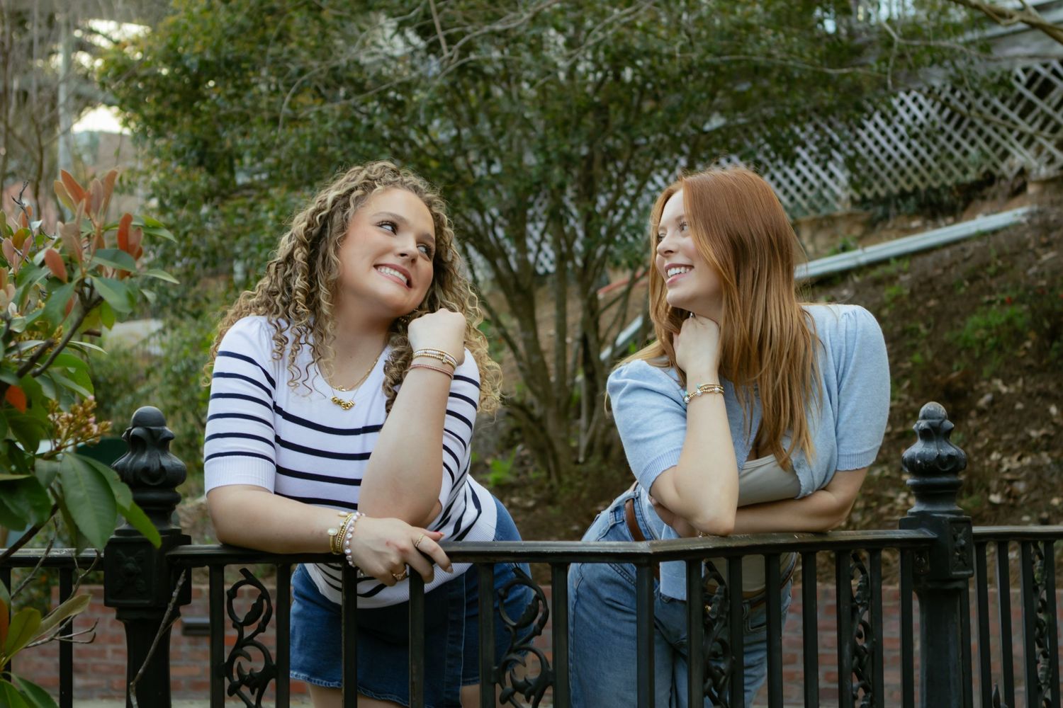 Two young women leaning on iron fence laughing outdoors among green foliage.