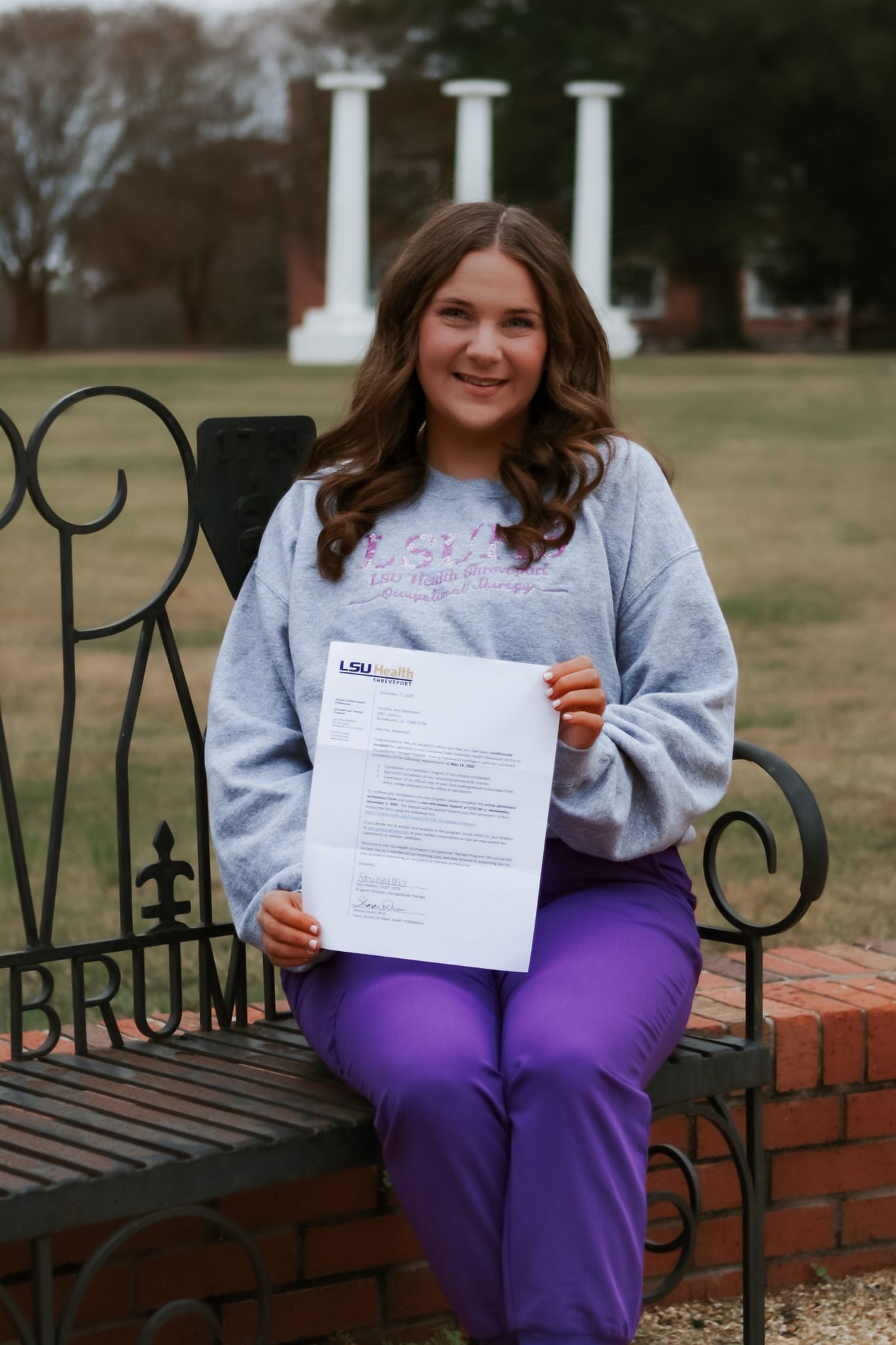 Person sitting on a brick wall bench wearing purple pants and holding paperwork.