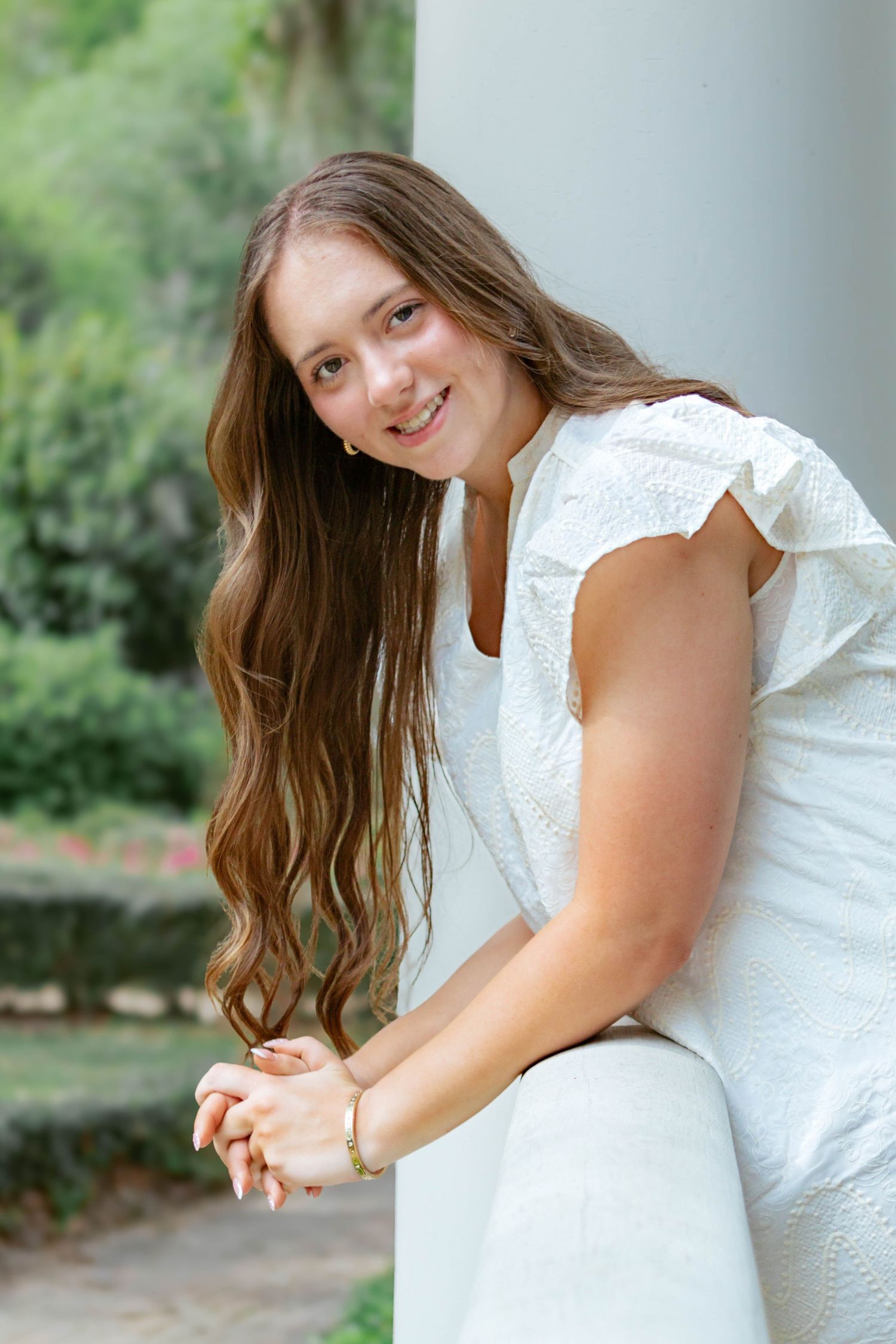 Smiling young woman with long wavy hair in white ruffled top leaning on white column outdoors.