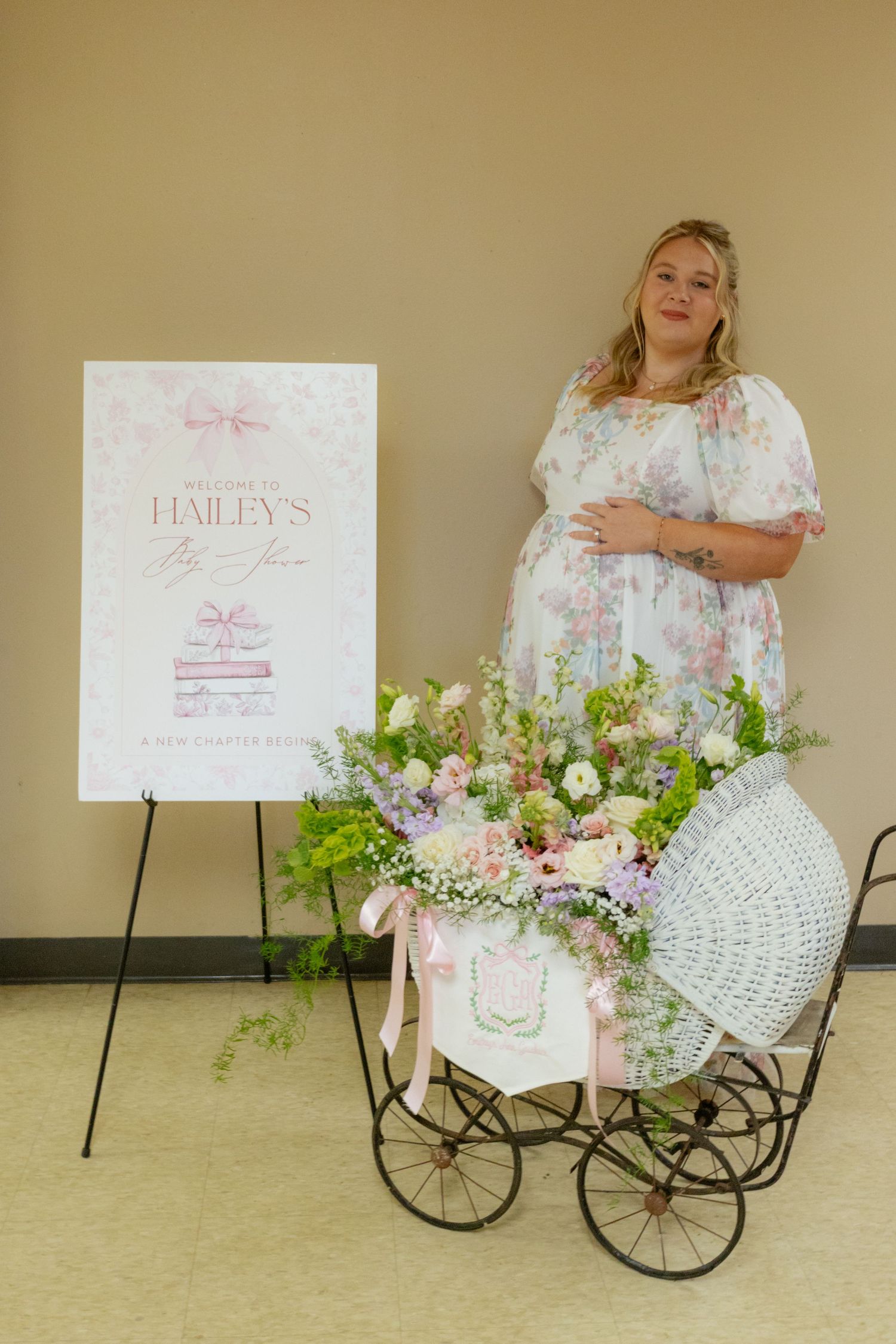 Pregnant woman in floral dress standing beside vintage wicker pram at Hailey baby shower event.
