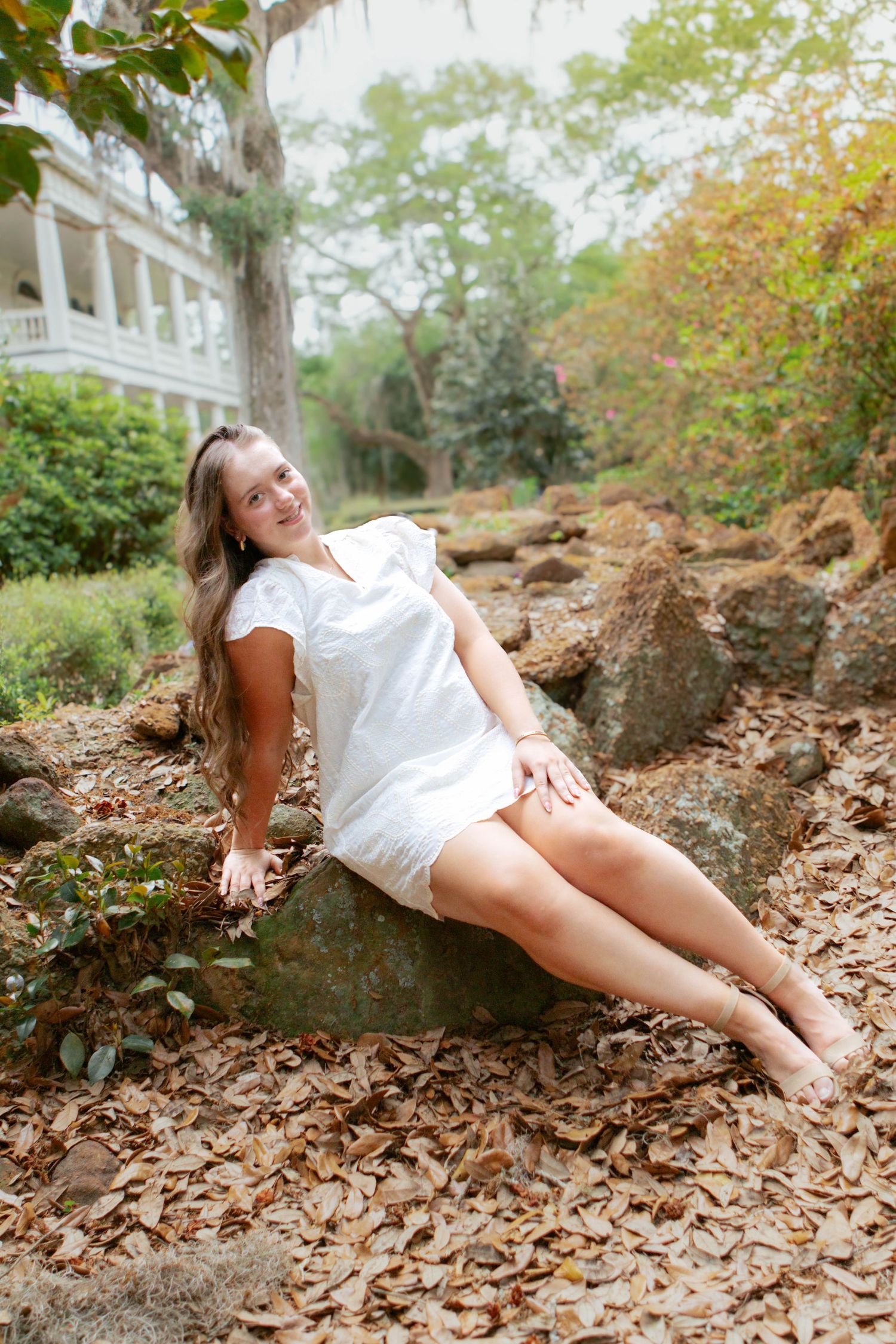 Young woman in white dress sitting on rocks surrounded by autumn leaves in a lush garden setting.