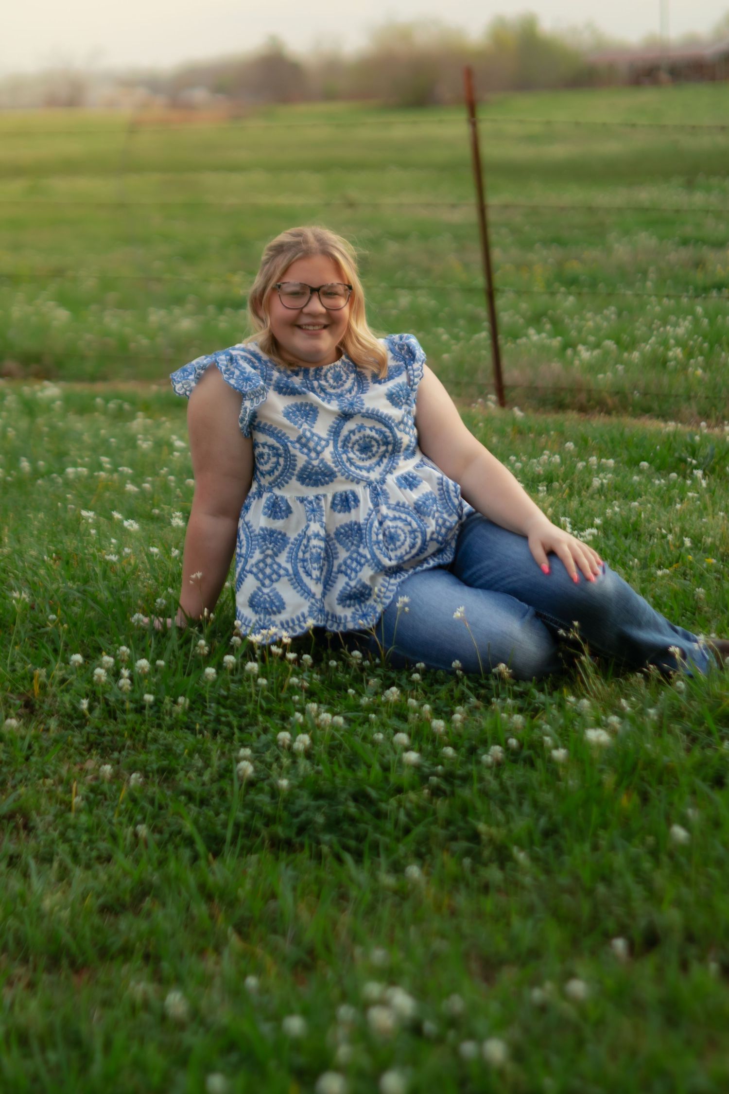Someone wearing a blue and white floral dress and jeans sits in a green field at sunset.