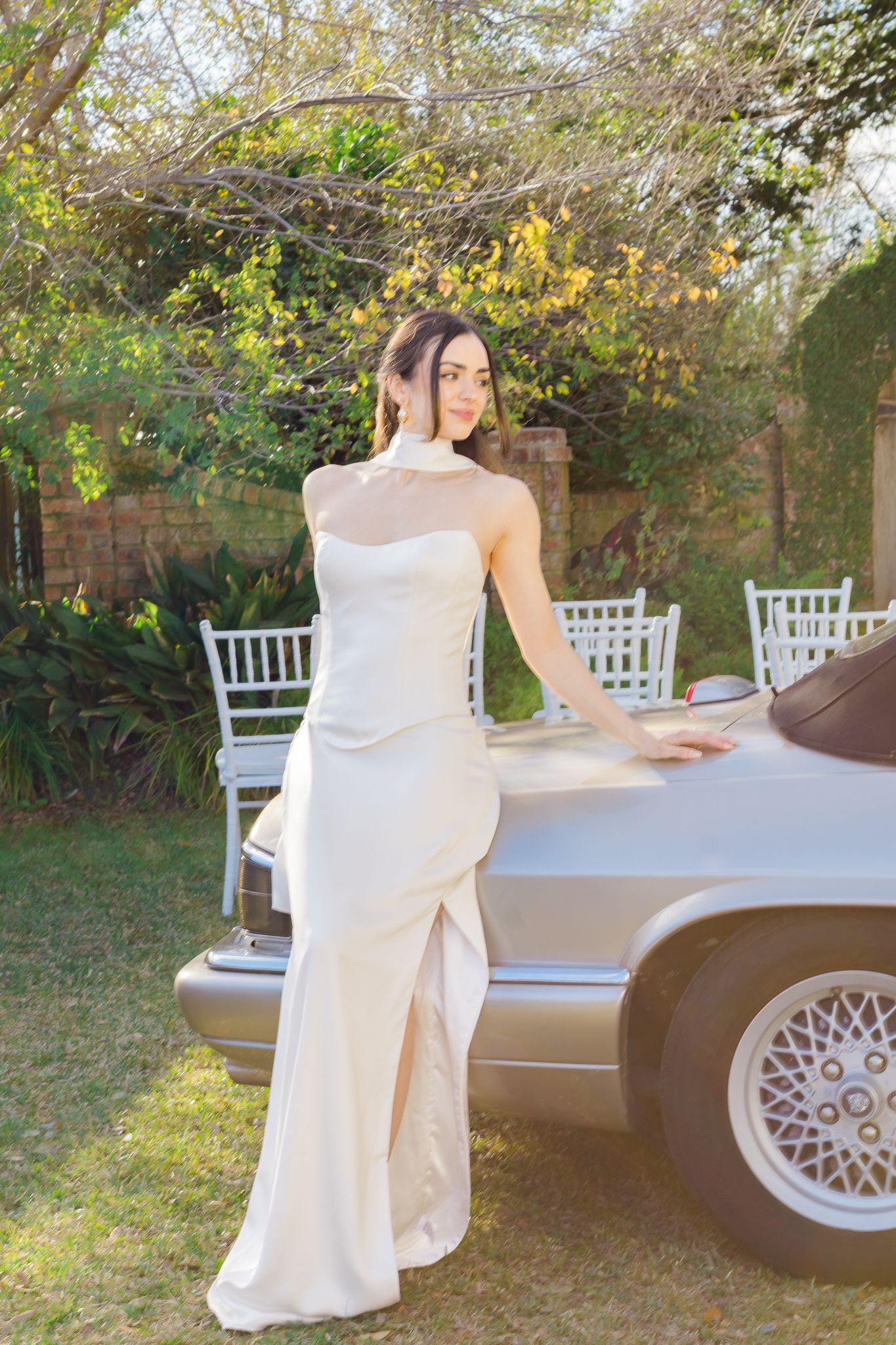 Elegant bride in a sleek ivory halter gown leaning against a silver convertible car in a lush garden setting.