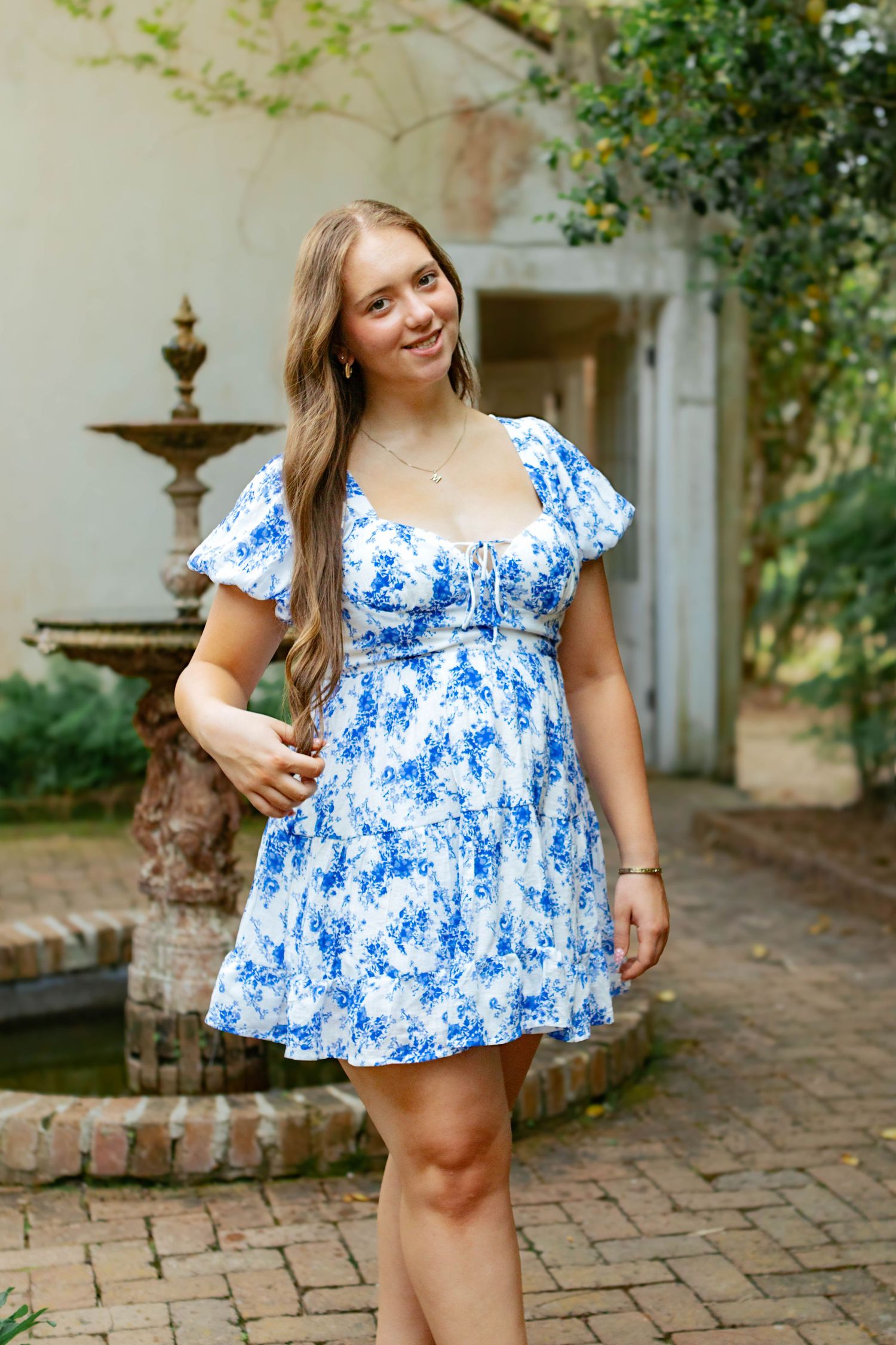 Young woman in blue floral mini dress posing near stone fountain in courtyard garden setting.