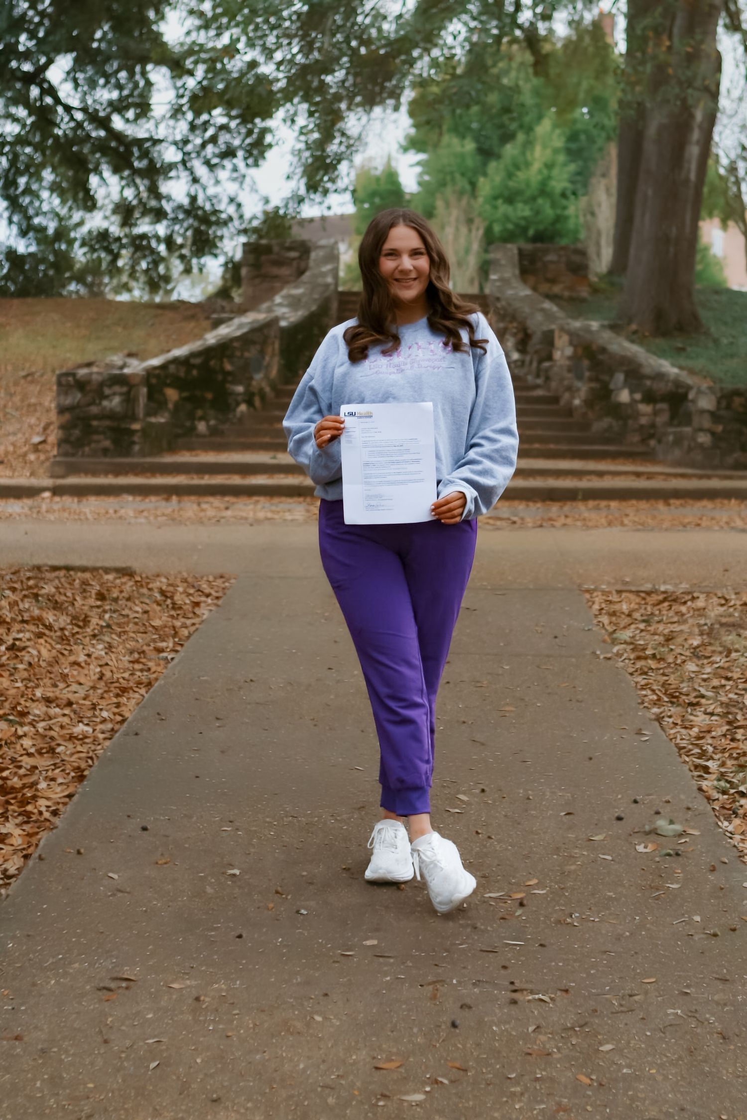 Person in purple pants and light blue sweater walking on a leaf-covered path holding a document.