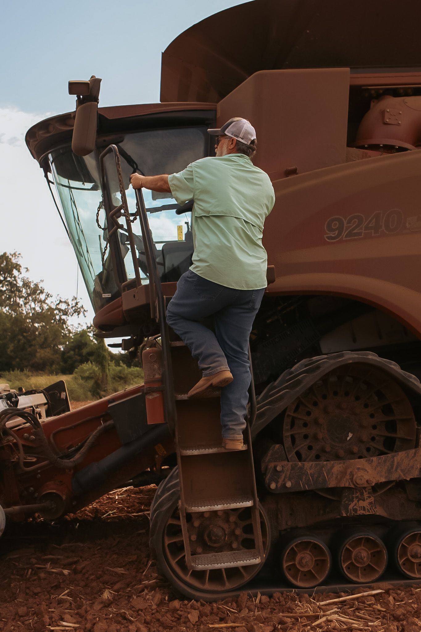 Farmer in green shirt and jeans climbing ladder to enter cab of large red combine harvester.