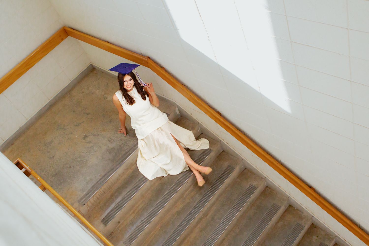 Graduate in cap and white dress sitting on stairs viewed from above.