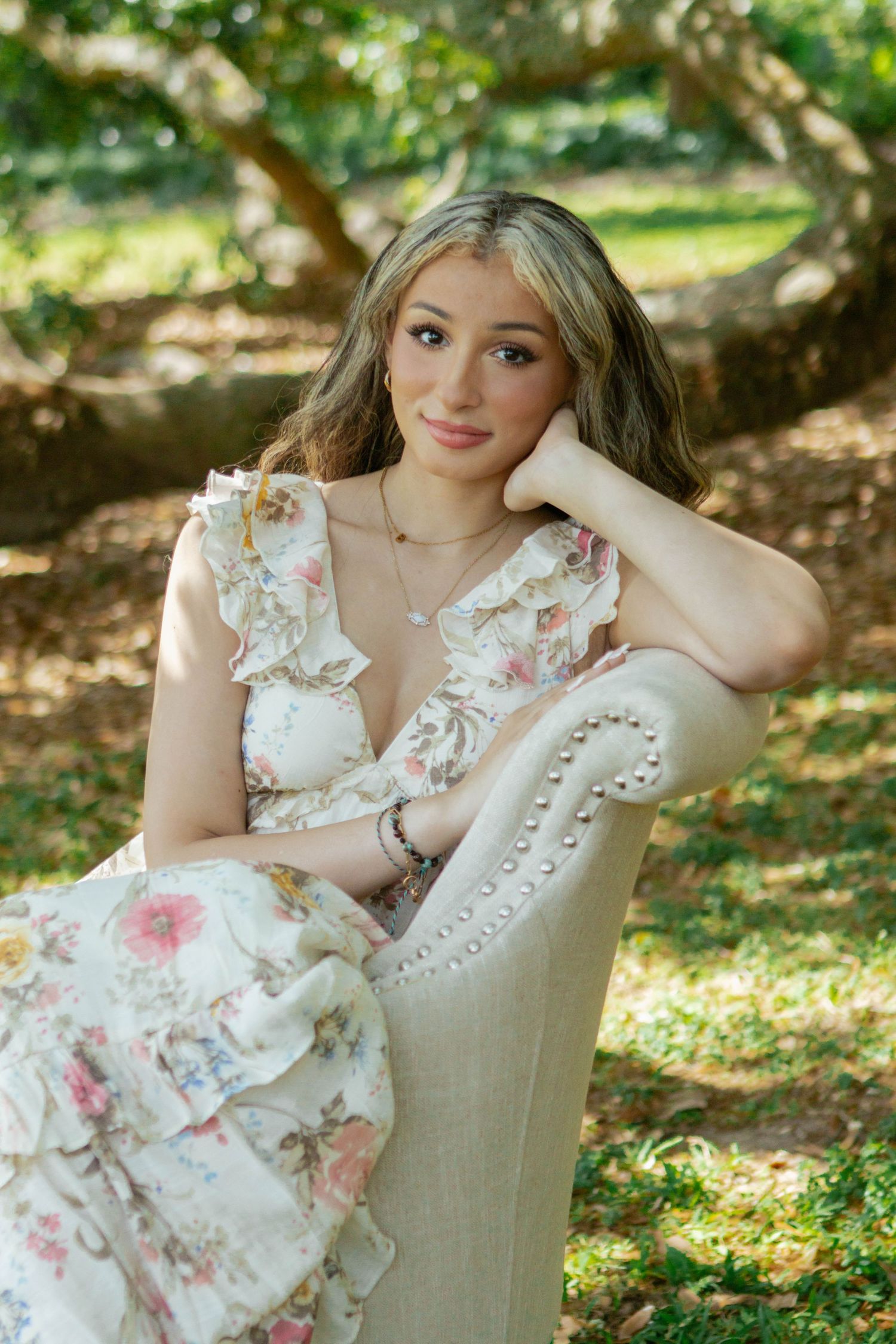 Young woman in floral ruffled dress sitting gracefully in an upholstered chair outdoors in a garden.