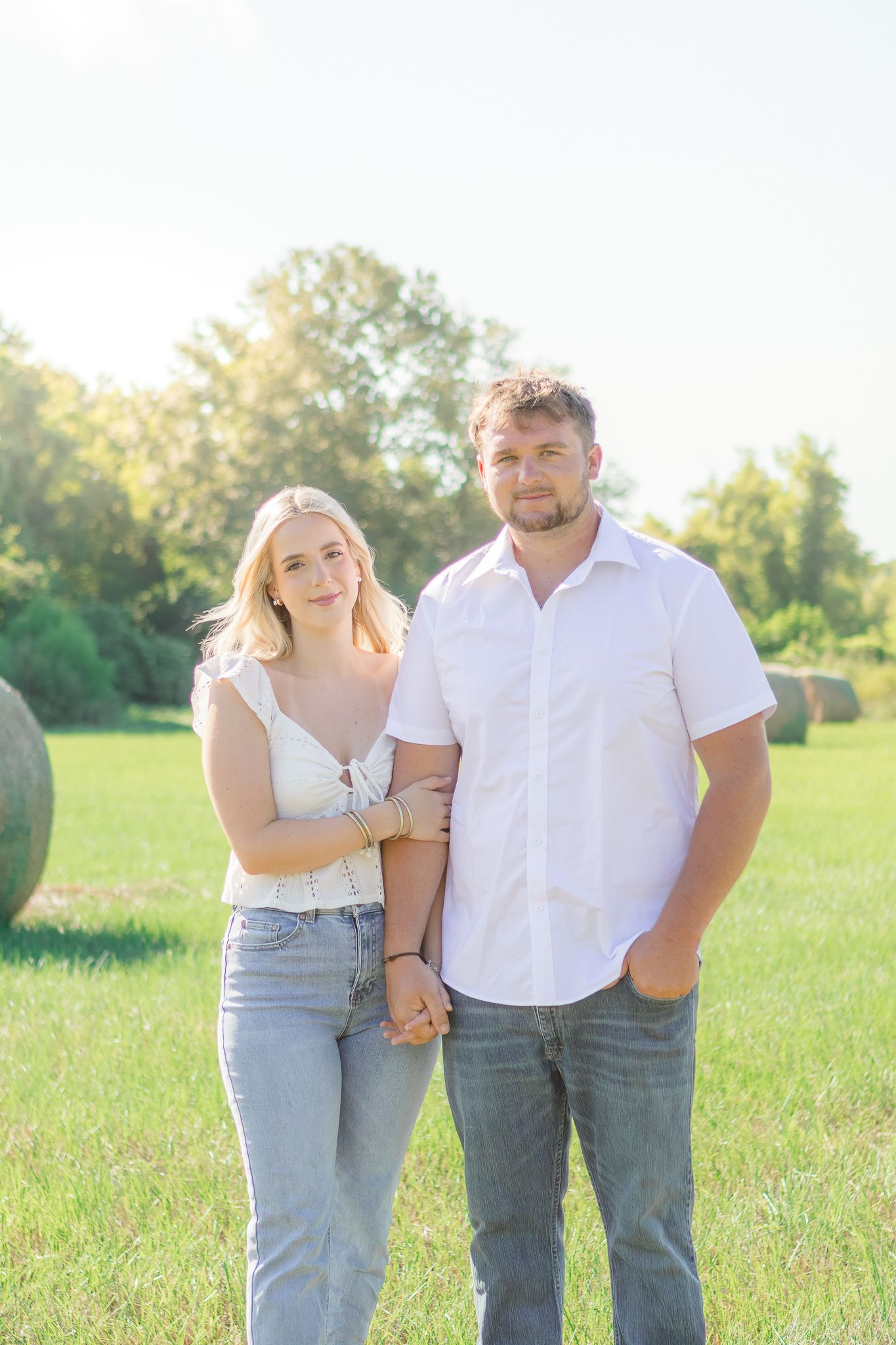 A couple holds hands in a sunny green field with hay bales, woman in white top and jeans, man in white shirt.