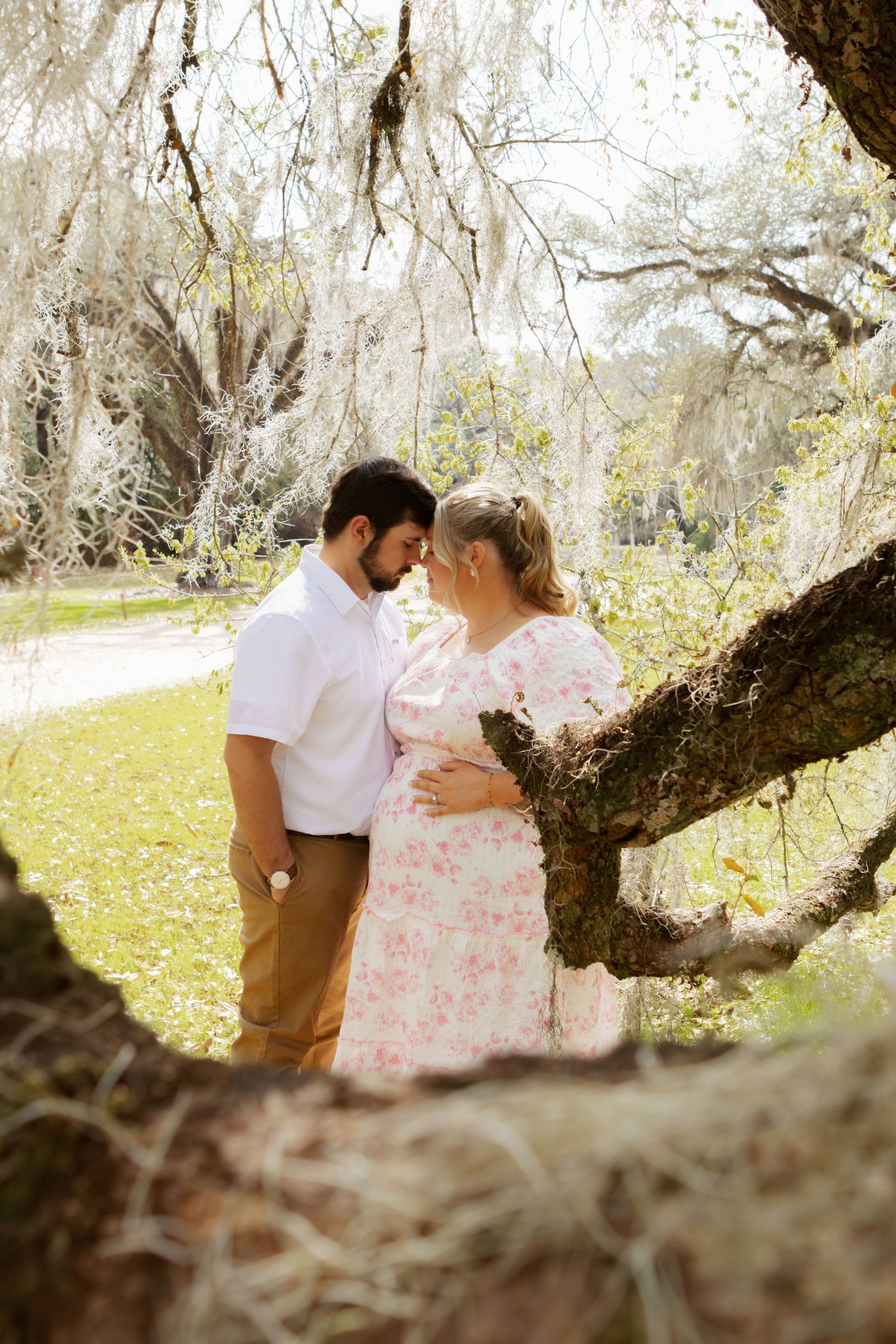 Romantic couple embraces under Spanish moss draped trees in a sunlit Southern garden setting.