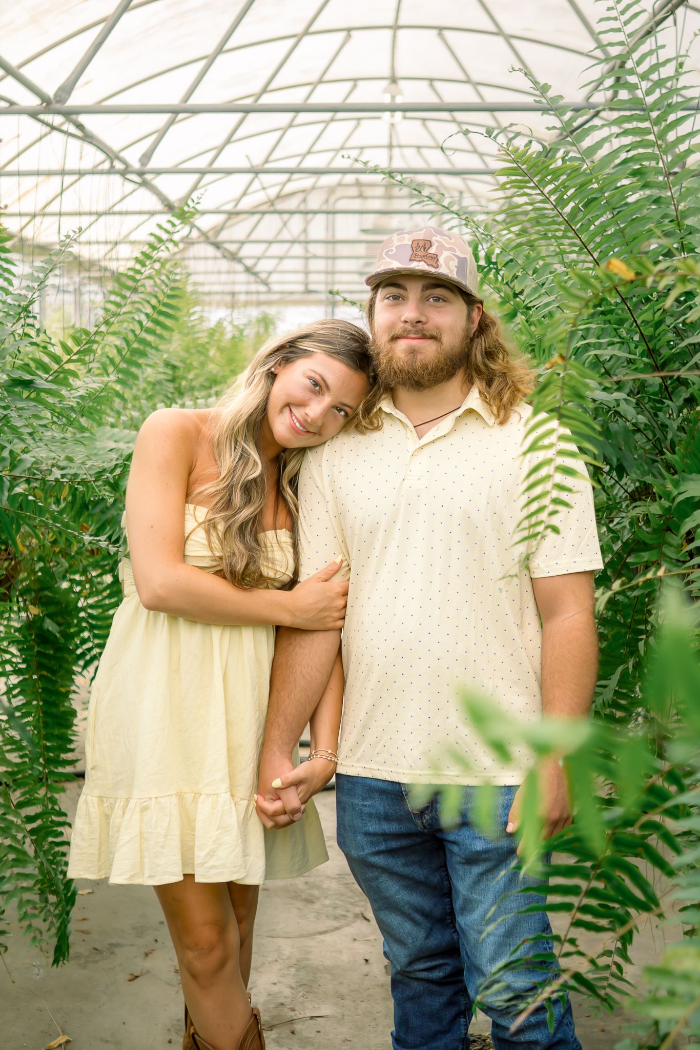 Smiling couple posing together among lush ferns inside a greenhouse, holding hands warmly.