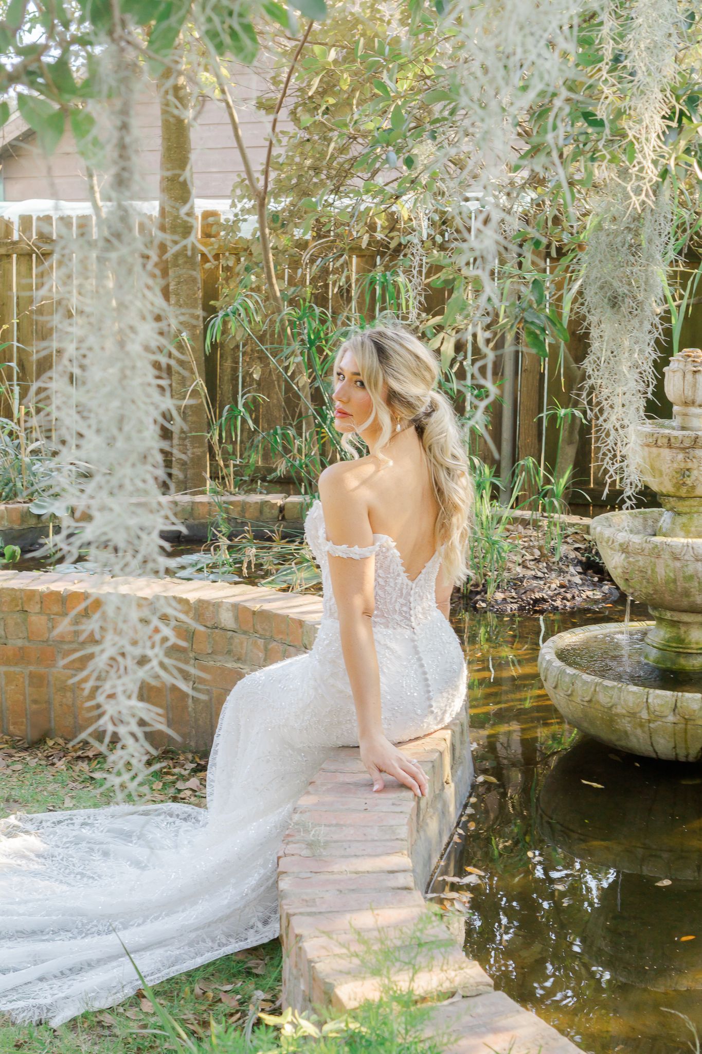 Bride in off-shoulder lace wedding gown sitting by garden pond with Spanish moss.