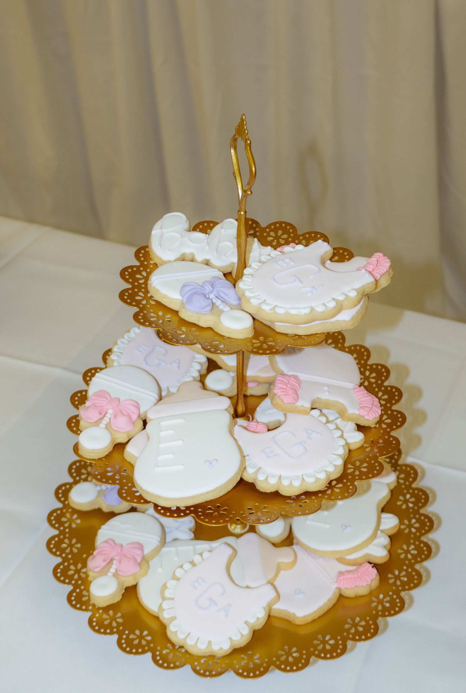 Three-tier gold stand displaying white and pink frosted baby shower cookies on a white tablecloth.