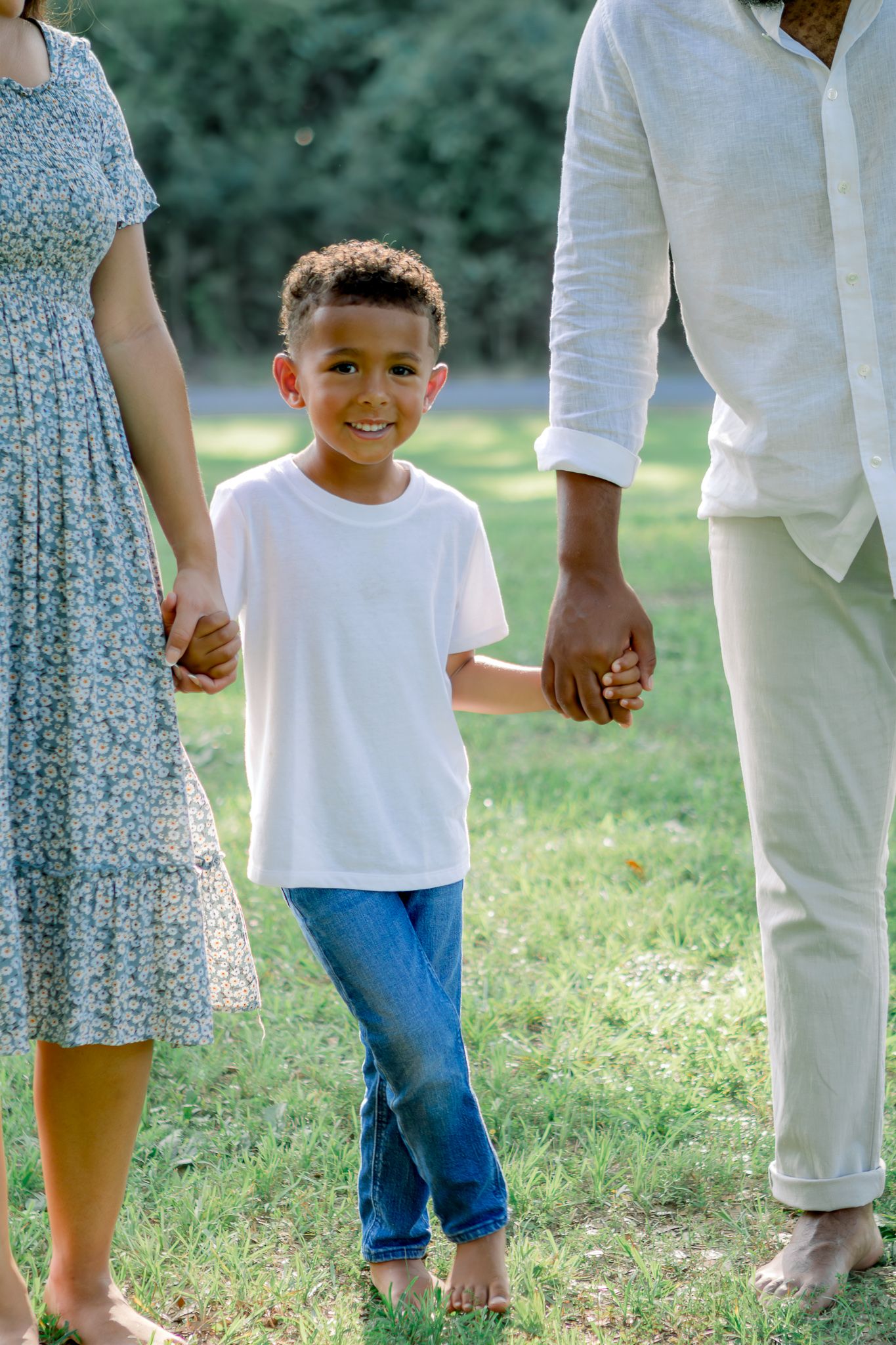 A smiling boy in white t-shirt and jeans holds hands with parents while walking barefoot on green grass outdoors.