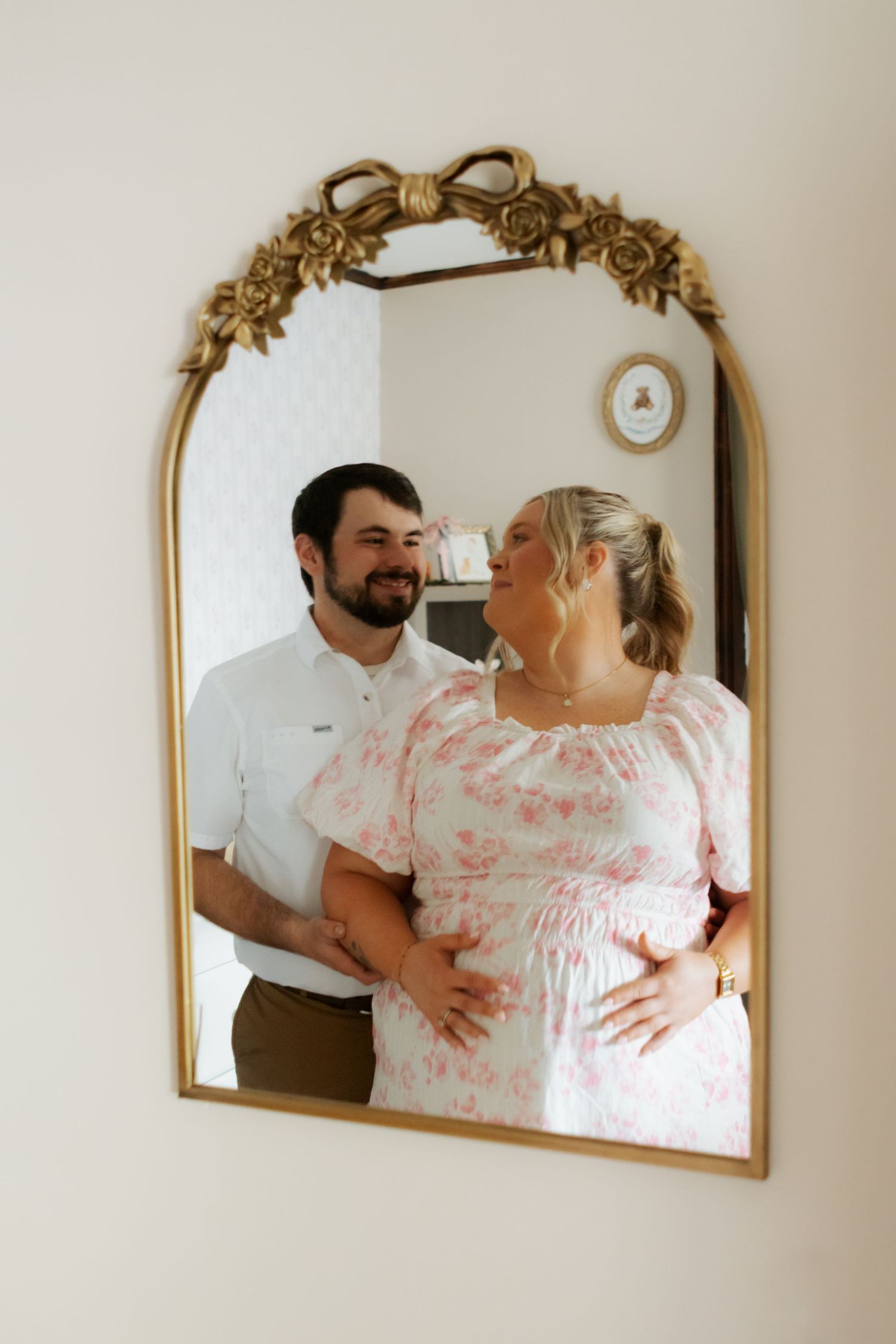 Pregnant couple smiling at each other in ornate gold mirror reflection, woman in pink floral dress with hands on baby bump.