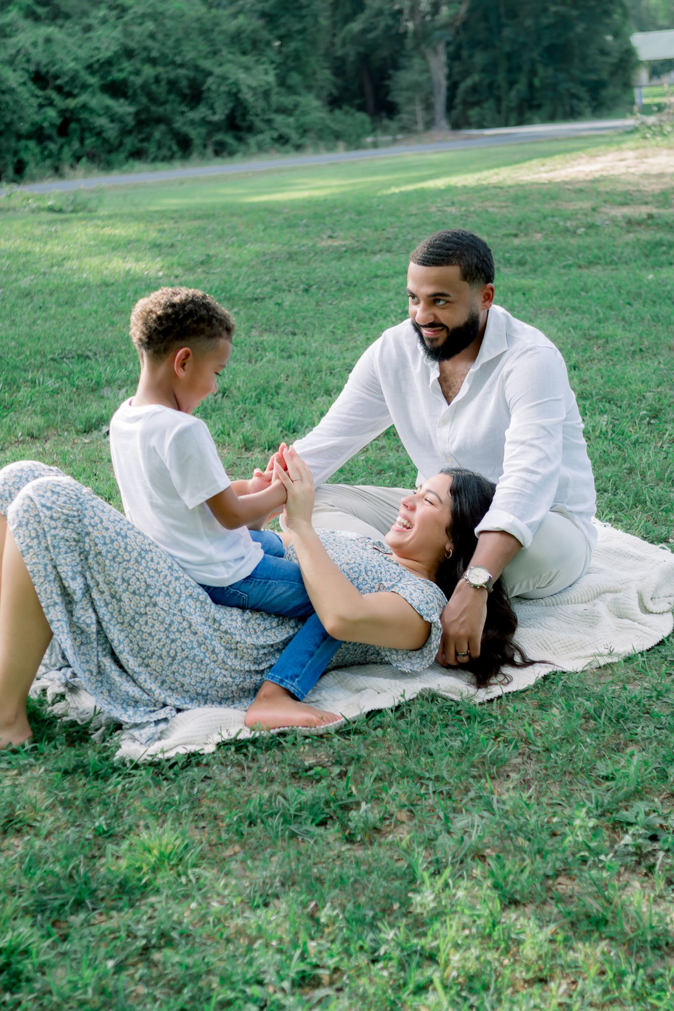 Family of three enjoying a picnic on green grass, with mom lying down laughing as dad and child hold hands.