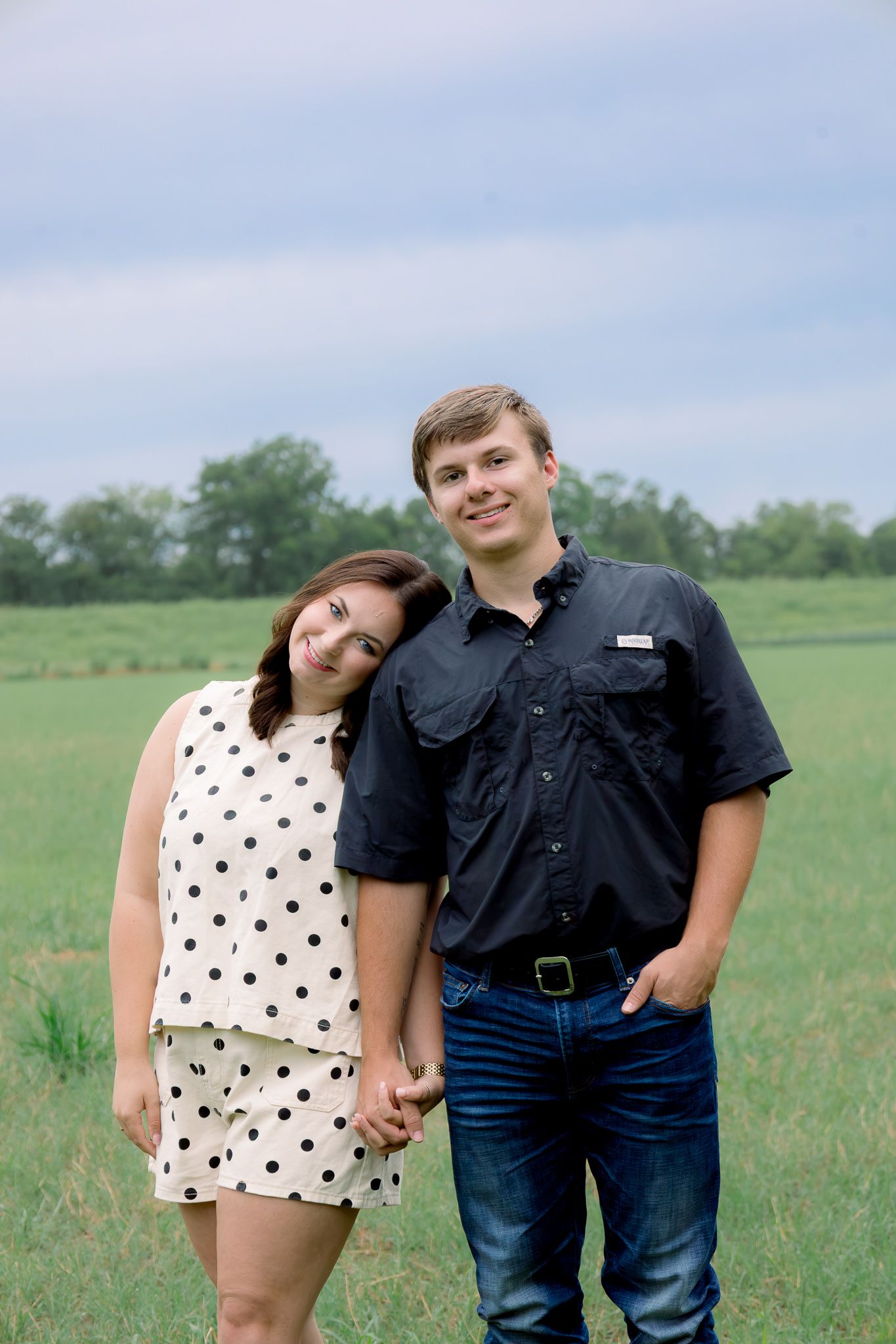 Young couple holding hands in a green field, woman in polka dot dress leaning on mans shoulder.