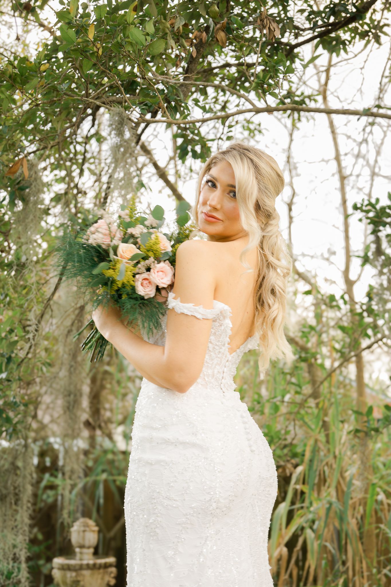 Blonde bride in an off-shoulder lace wedding gown holding blush roses while posing among lush garden trees.