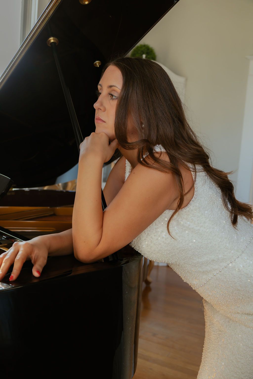Brunette bride in white sequin gown leans pensively against black grand piano indoors.