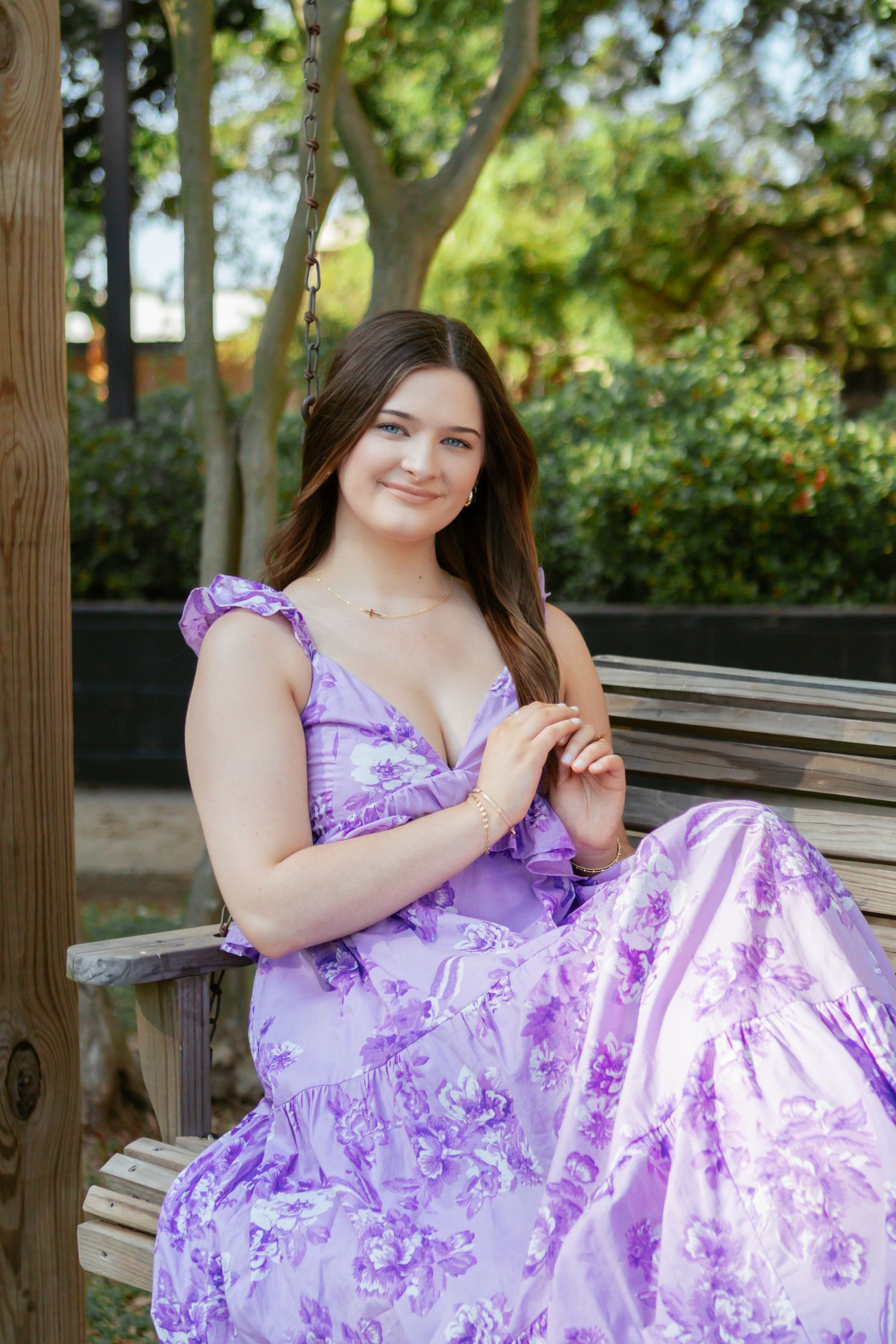 Young woman in purple floral maxi dress sitting on wooden swing bench in outdoor garden setting.