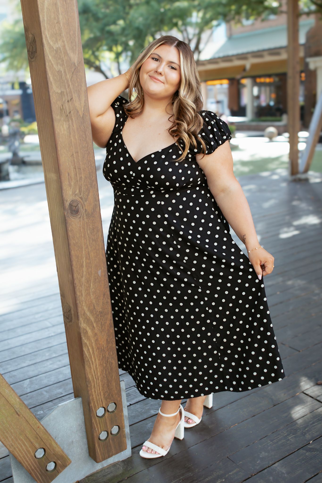 Young woman in black and white polka dot midi dress posing against a wooden post on an outdoor patio.