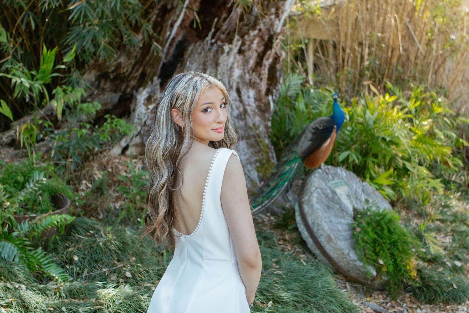 Blonde woman in white dress smiles over shoulder with colorful peacock in garden background.