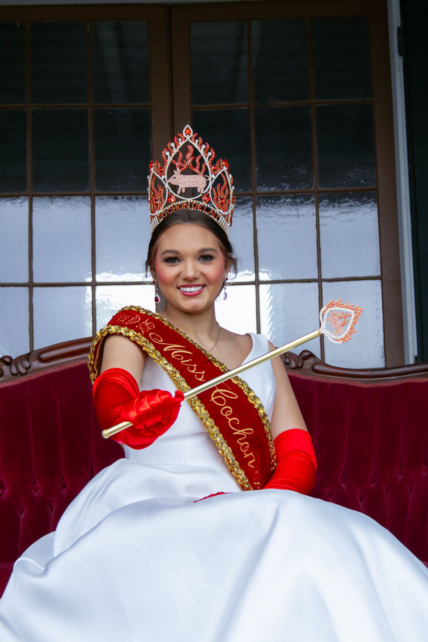 Miss Cochon beauty queen in white gown, red crown, sash and gloves holding decorative wand on throne.