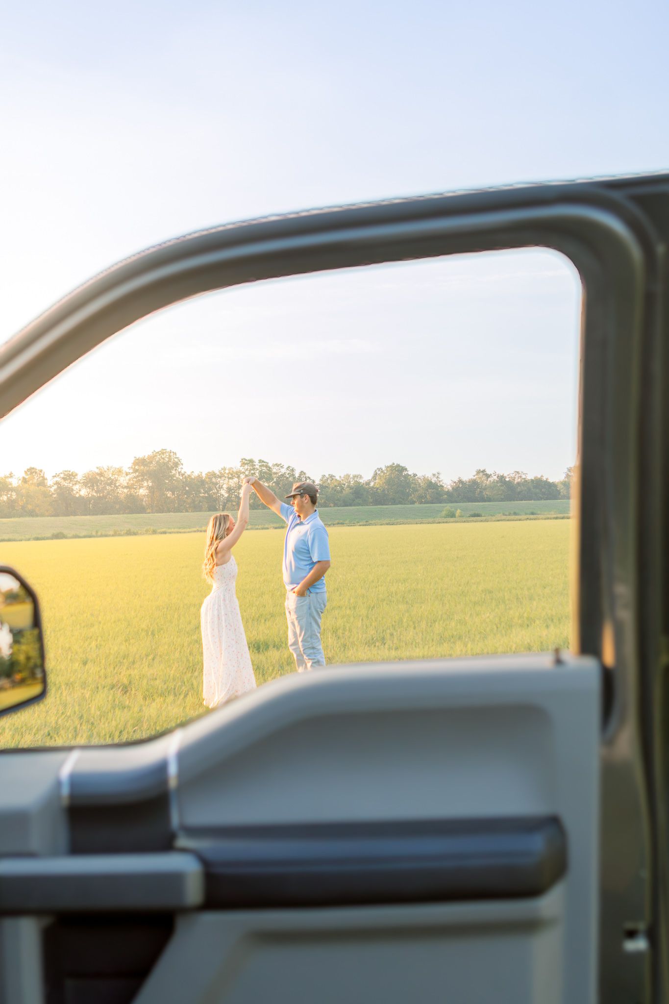 Couple dancing in a green field at golden hour, framed by a truck door in the foreground.