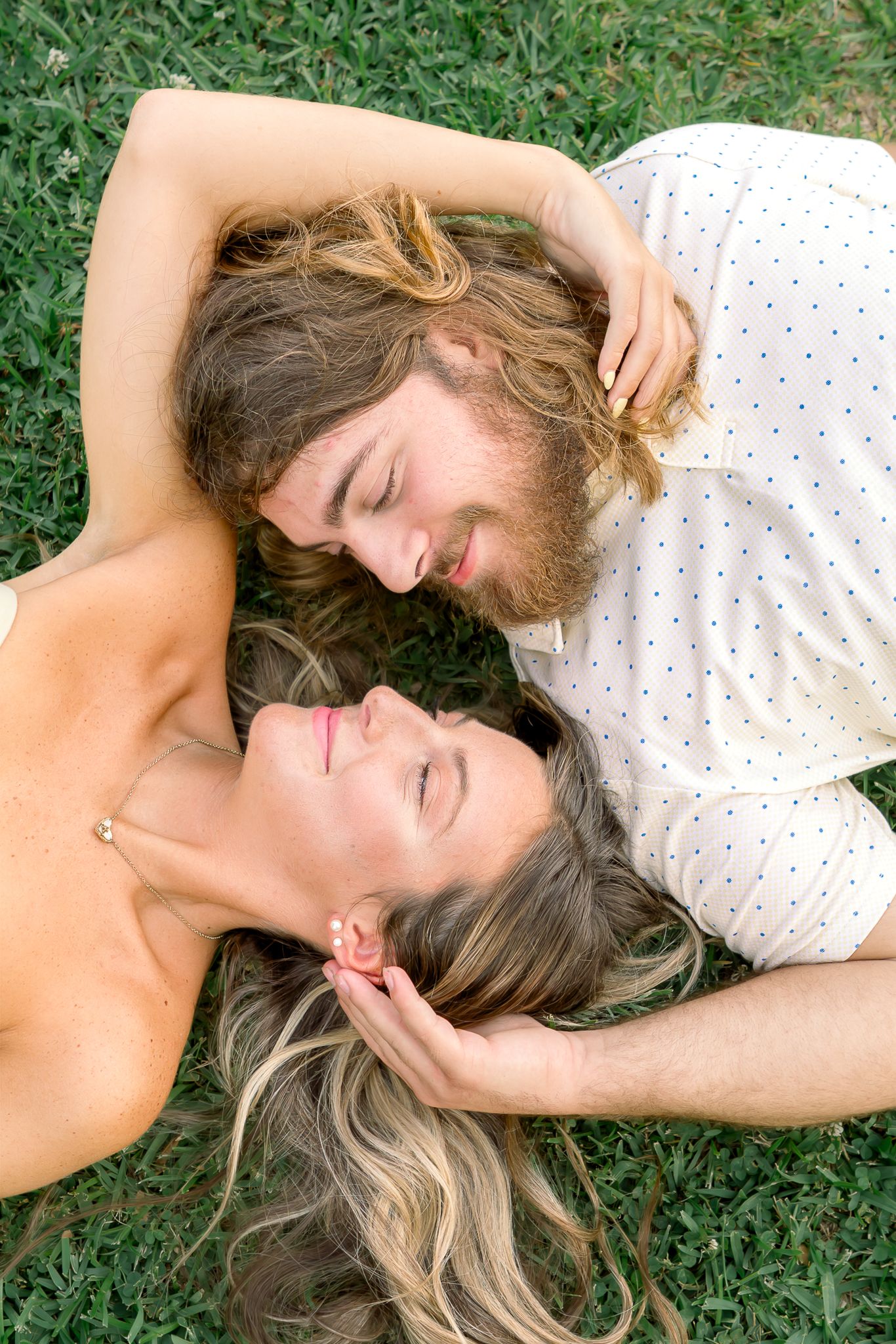 Couple lying head-to-head on green grass, smiling softly with eyes closed in a peaceful outdoor setting.