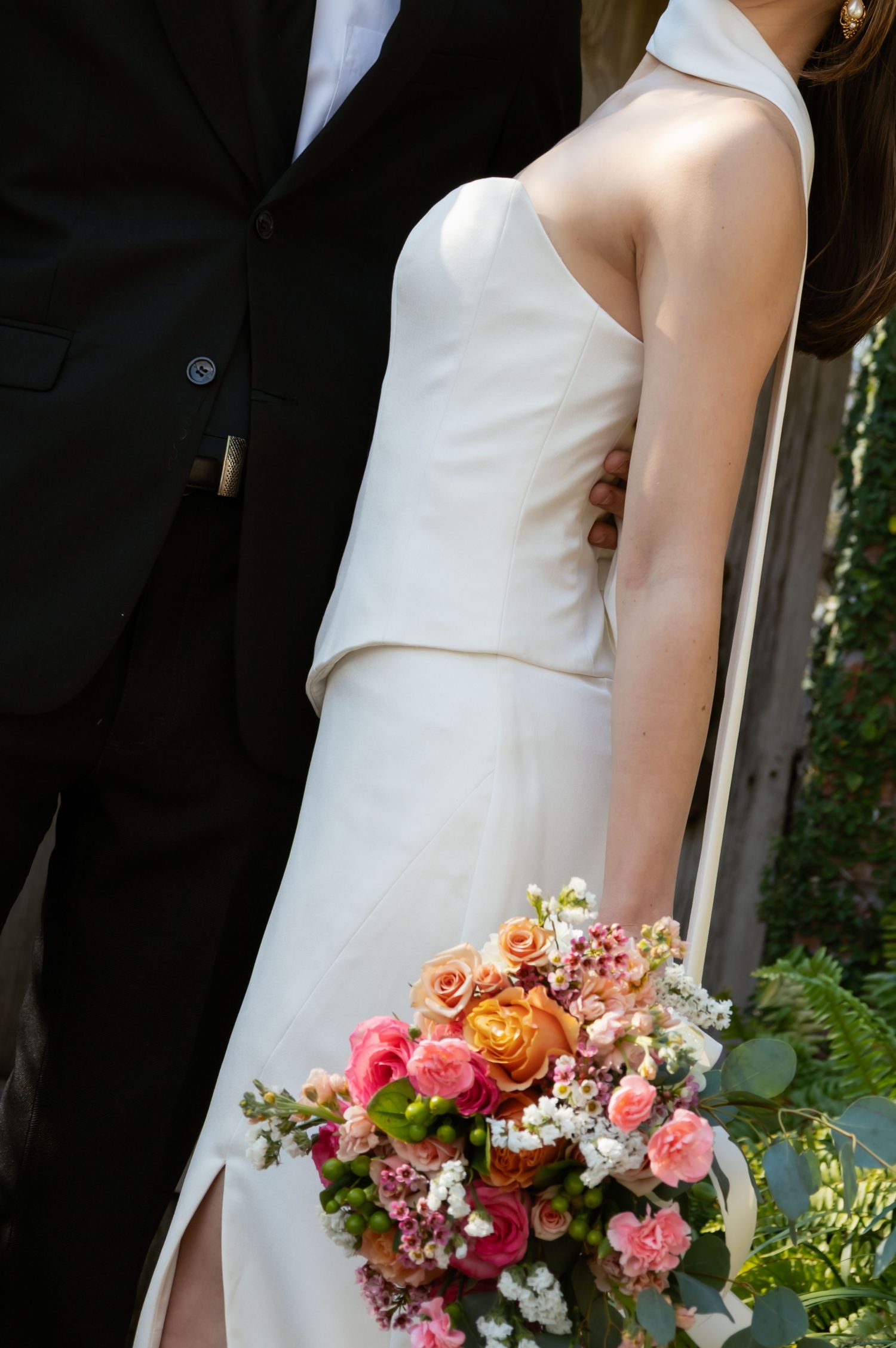 Bride in sleek white strapless gown holding a vibrant pink and orange floral bouquet beside a suited groom.