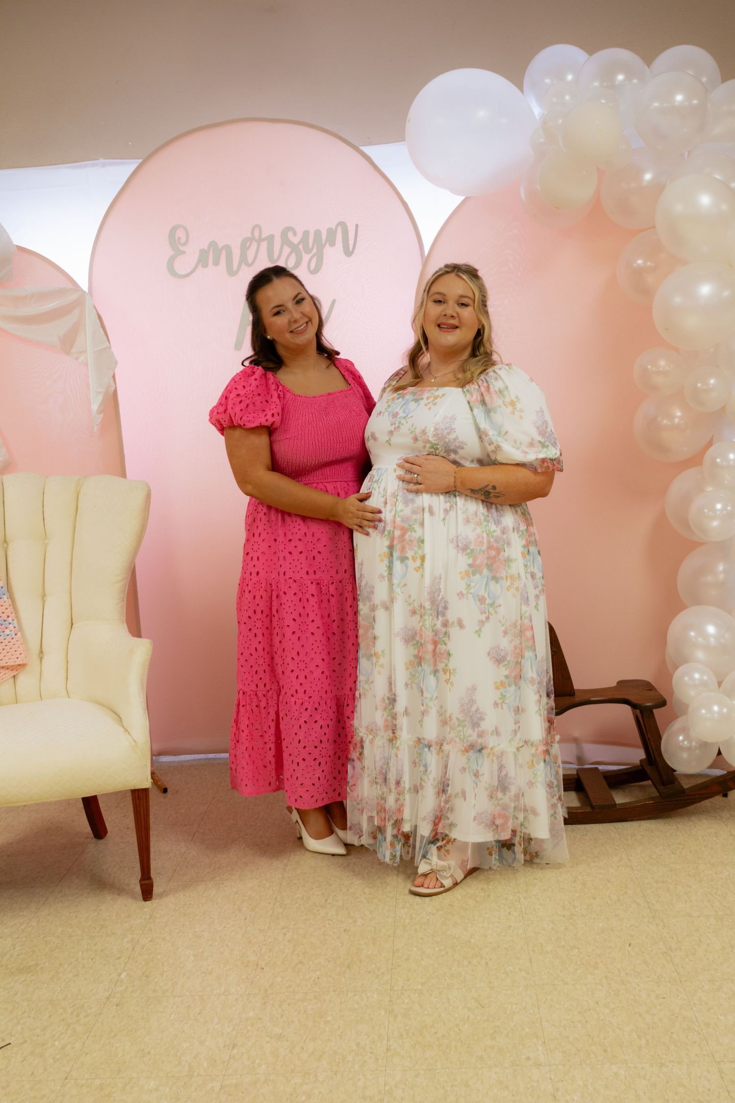 Two women posing at baby shower with pink balloon arch and Emersyn name sign backdrop.