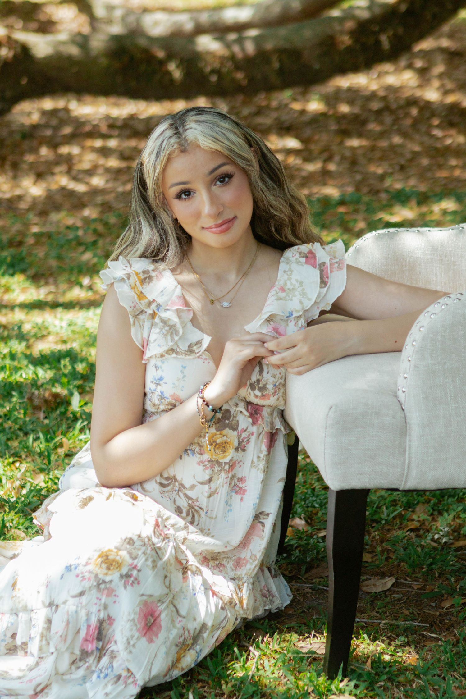 Young woman in floral ruffle dress sitting on grass leaning against cream upholstered chair outdoors.