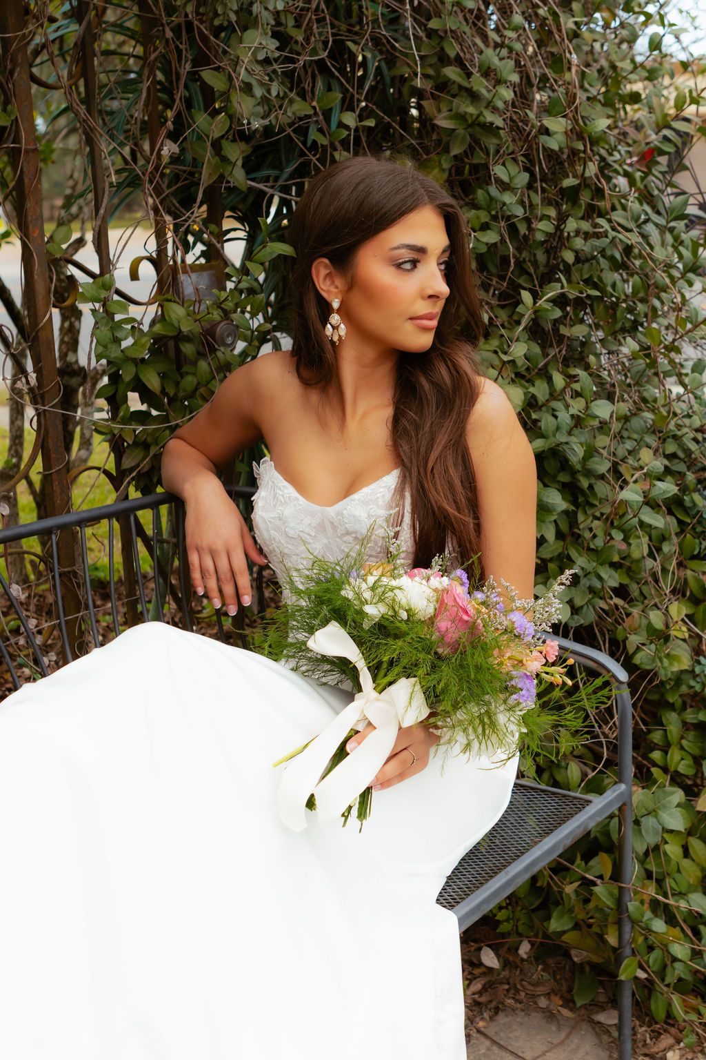 Brunette bride in a strapless lace gown seated outdoors holding a wildflower bouquet beneath a green ivy arch.